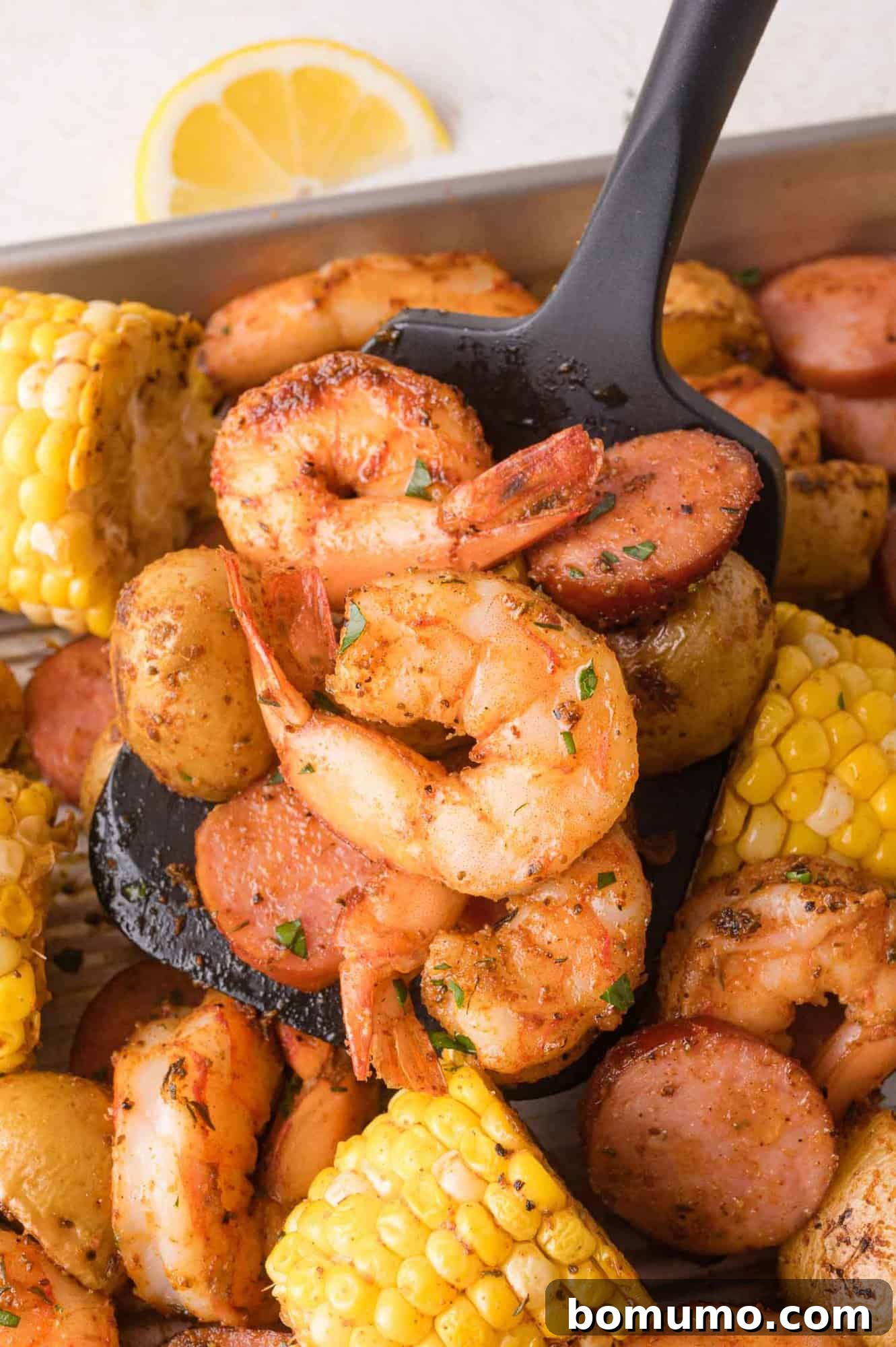 A black serving spoon holds seasoned shrimp, sliced sausage, potatoes, and corn from a tray of cooked seafood and vegetables. A lemon wedge is in the background.