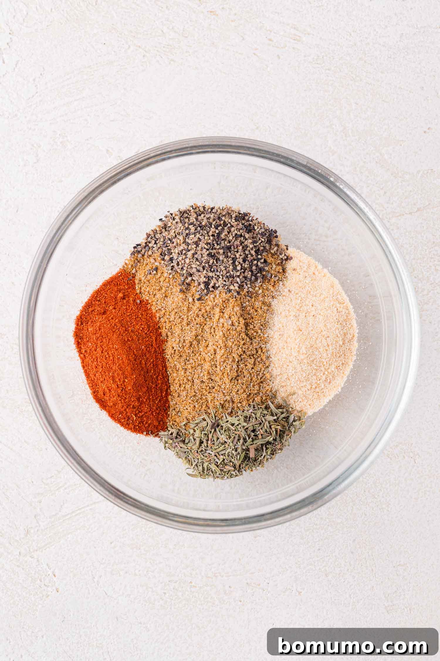 A glass bowl containing separate piles of dried spices, including paprika, black pepper, garlic powder, thyme, and brownish seasonings, on a light surface, ready to be mixed for a sheet pan shrimp boil.