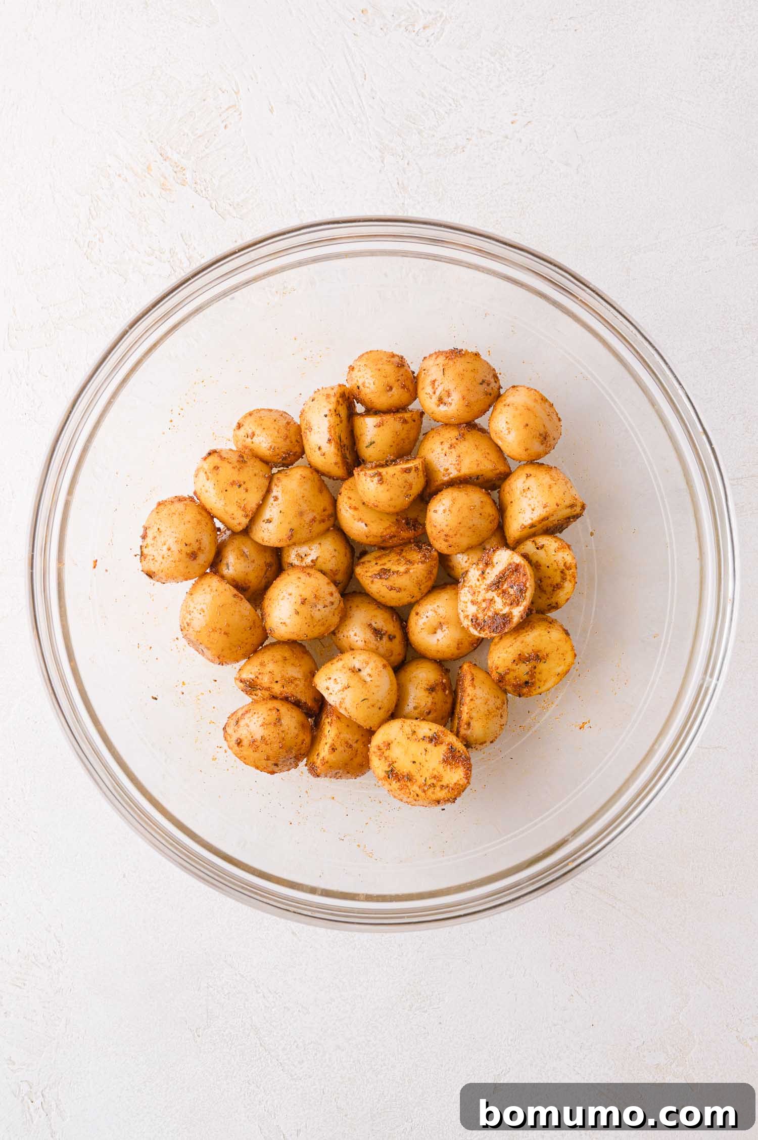 A glass bowl filled with halved baby potatoes, thoroughly coated in a vibrant seasoning blend, awaiting placement on the baking sheet for roasting.