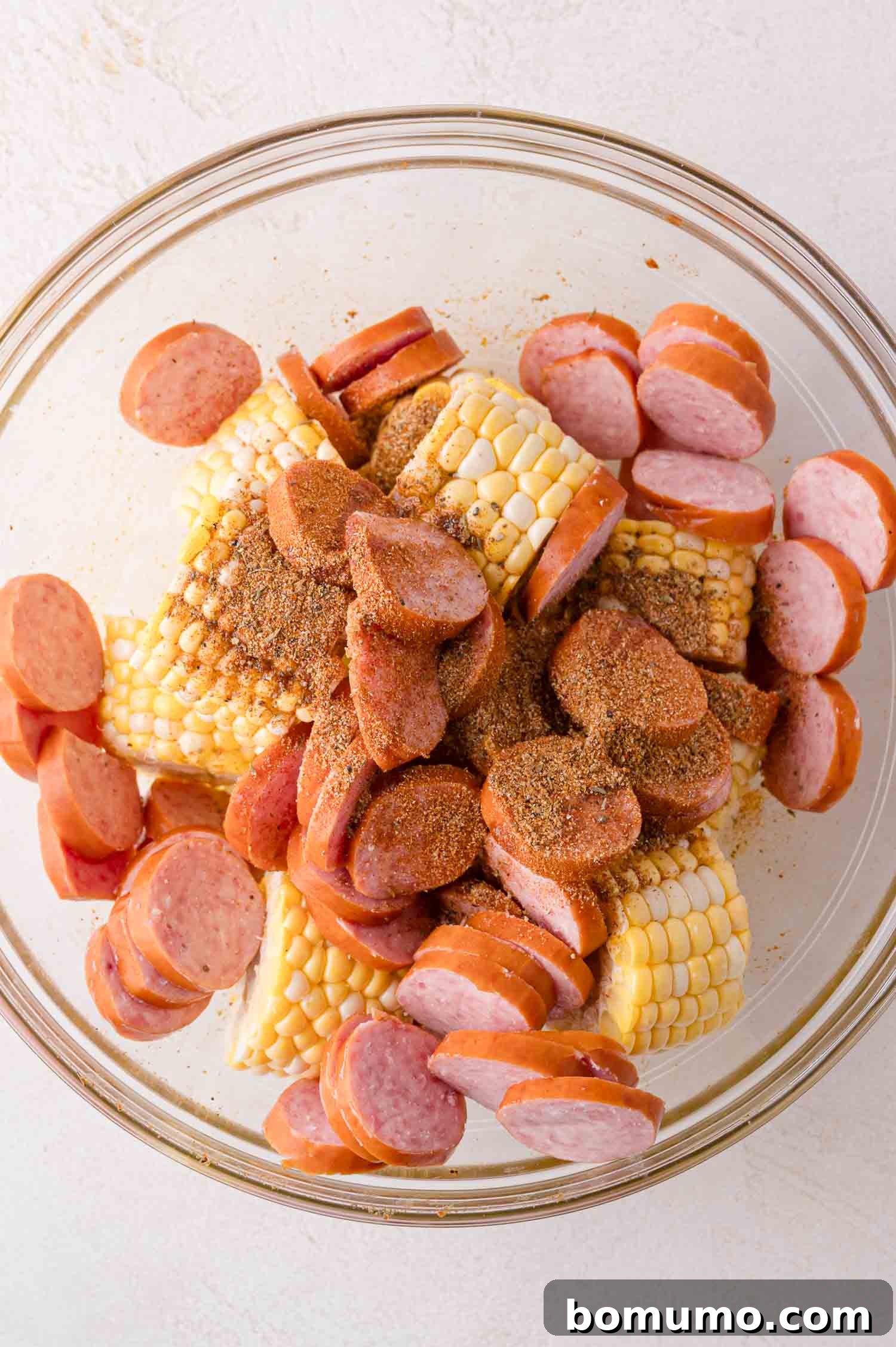 A glass bowl showcasing perfectly seasoned sliced sausage and vibrant corn on the cob pieces, ready to be added to the sheet pan for roasting.