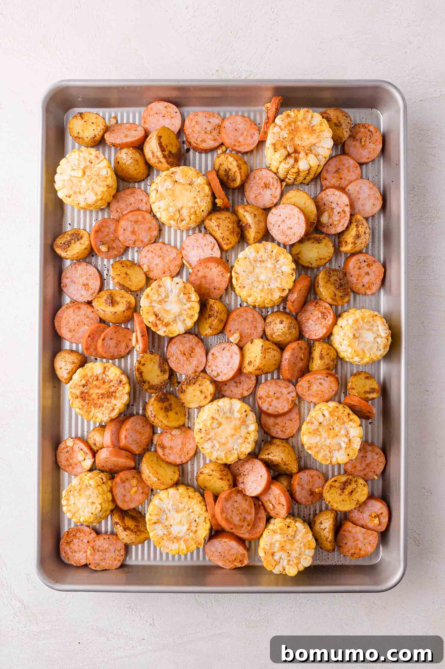 A metal baking tray with an artful arrangement of roasted baby potatoes, sliced smoked sausage, and corn on the cob rounds, all uniformly seasoned and ready for the next stage of cooking.