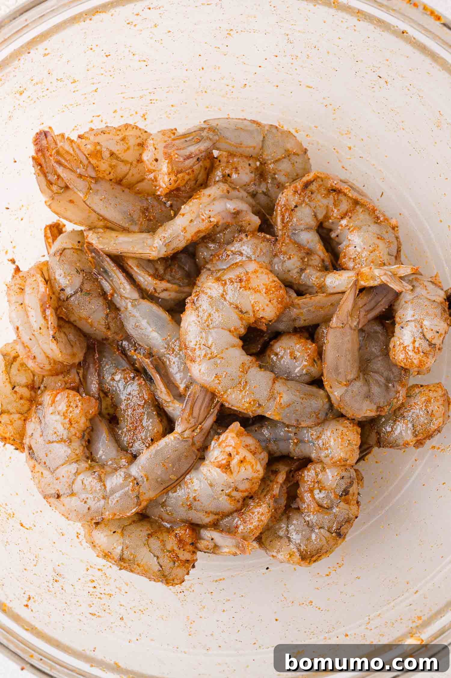 Raw shrimp, perfectly coated with a savory seasoning blend, resting in a clear glass bowl.
