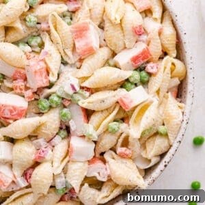 A close-up of a pasta salad with shell pasta, imitation crab meat, green peas, and diced red onion in a creamy dressing.