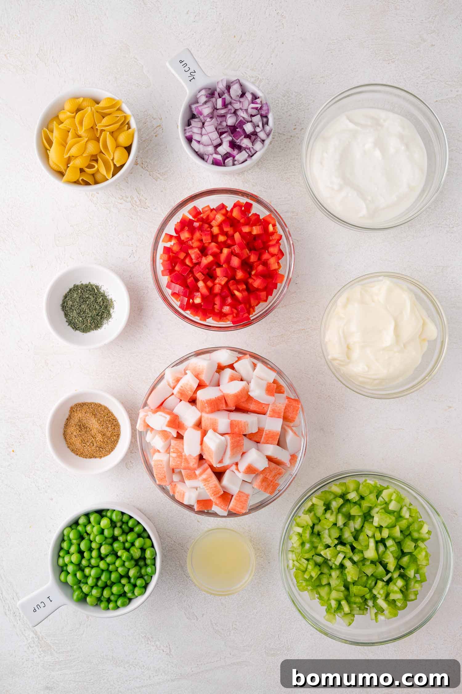 A selection of fresh ingredients neatly arranged in bowls, including uncooked pasta, finely chopped red onion, bell pepper, green peas, crisp celery, imitation crab flakes, and seasonings, along with lemon juice, yogurt, and mayonnaise for the dressing.