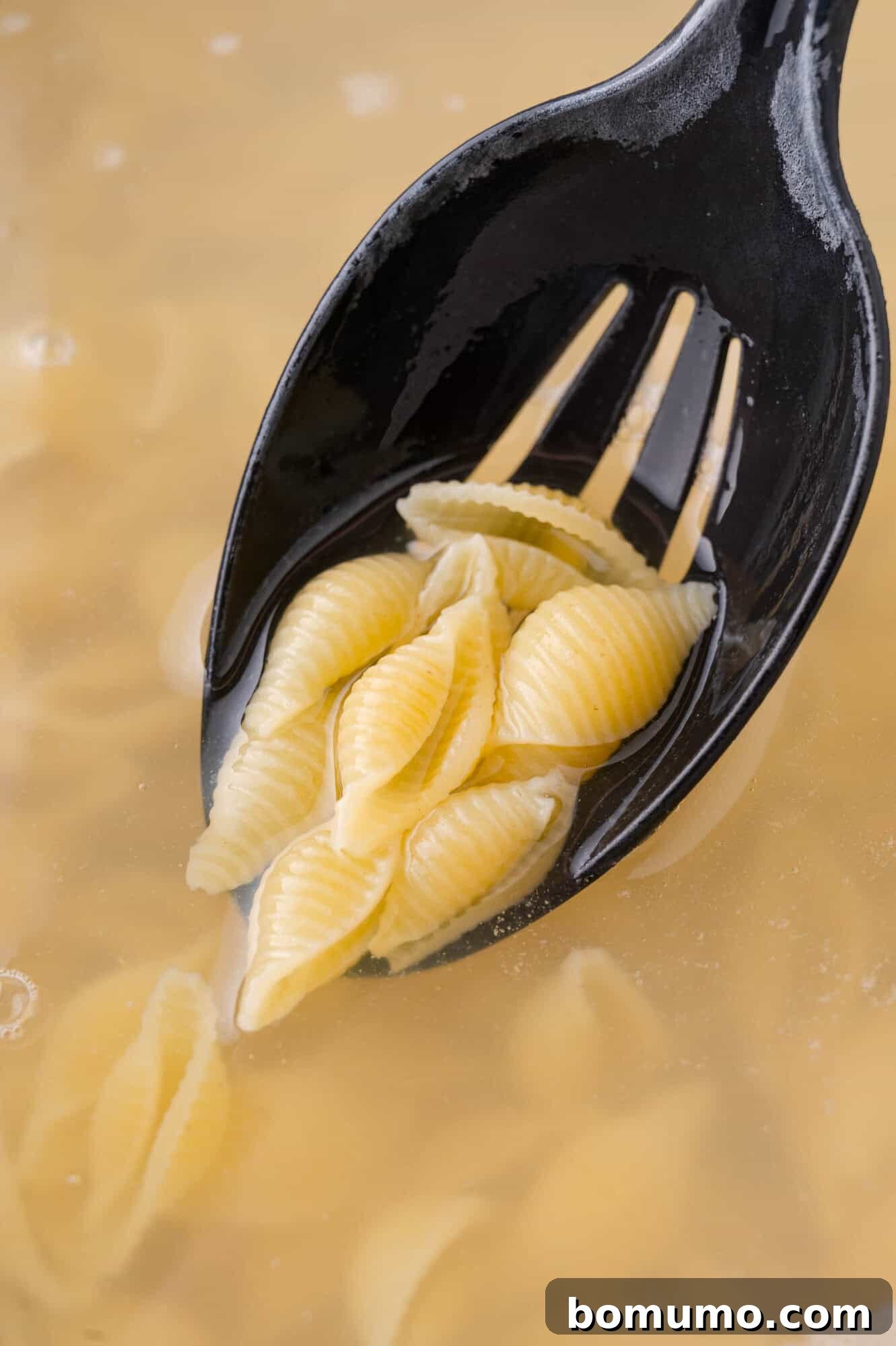 A black slotted spoon holds perfectly cooked shell pasta, rising steam from a pot of boiling water, illustrating the pasta preparation stage.