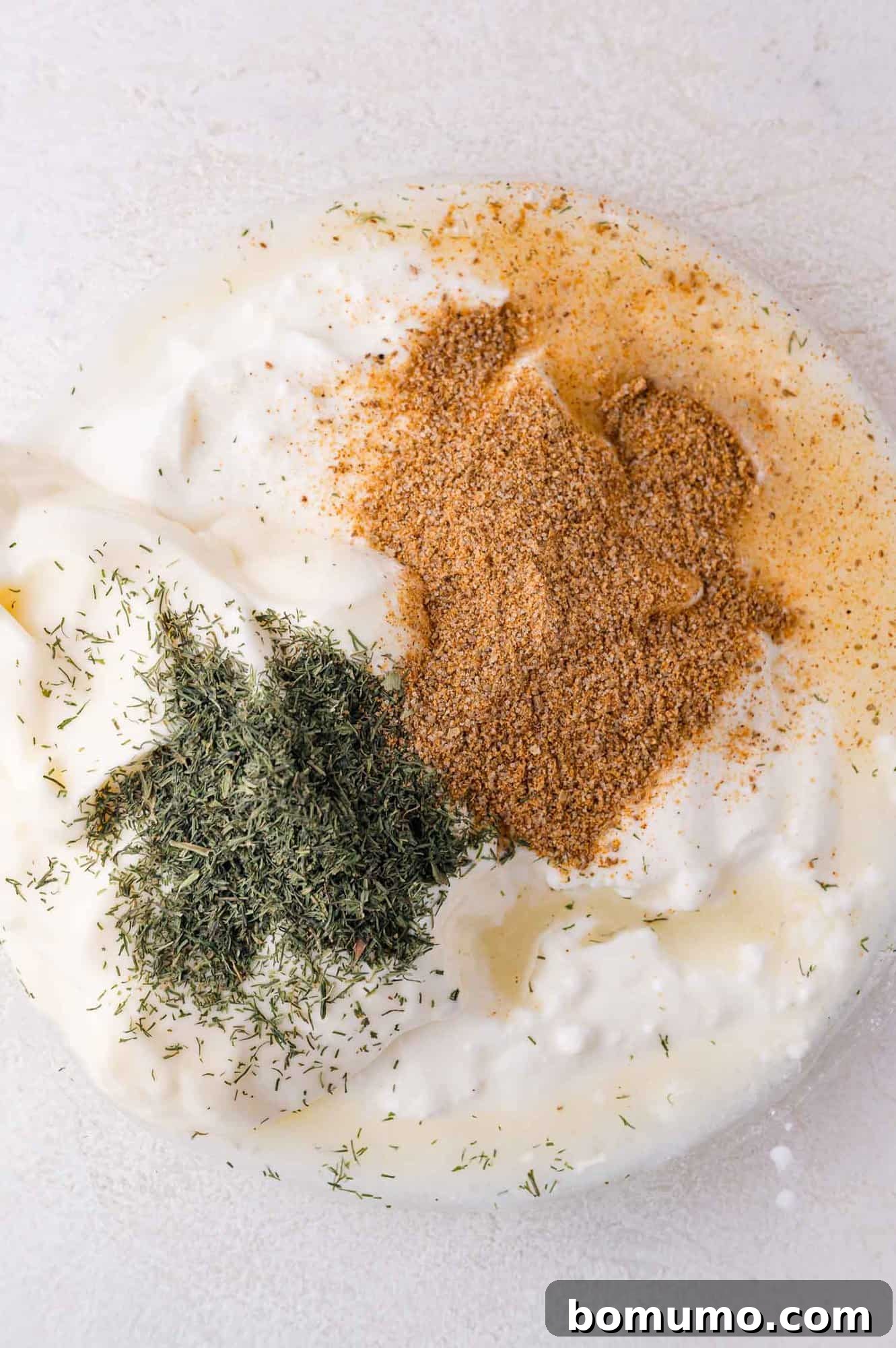A creamy white sauce in a bowl, generously sprinkled with dried dill and Old Bay seasoning, set against a light background, highlighting the key dressing ingredients.