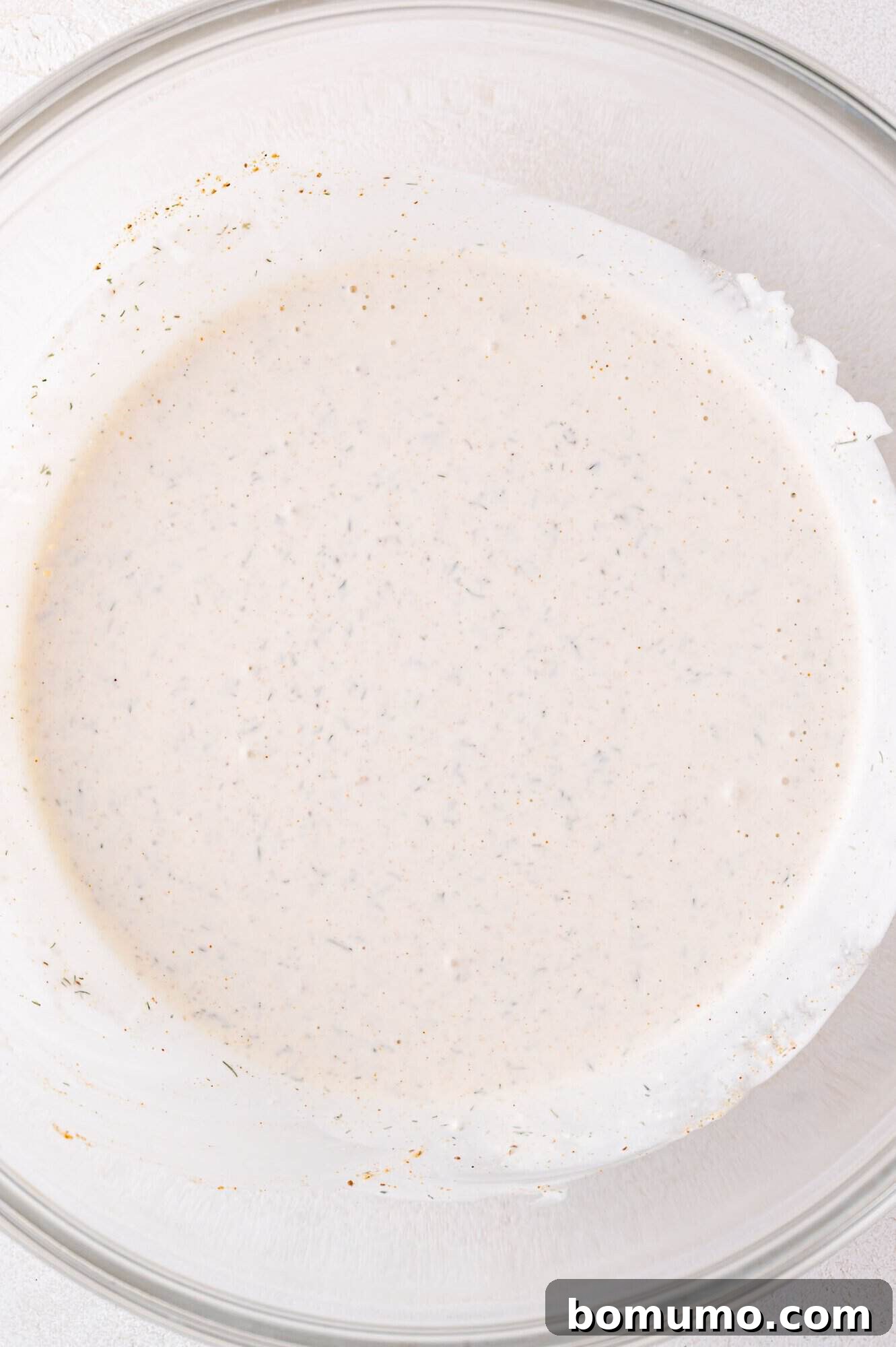 A clear glass bowl filled with a perfectly blended, pale creamy sauce, visibly speckled with aromatic herbs and spices, showcasing the prepared dressing.