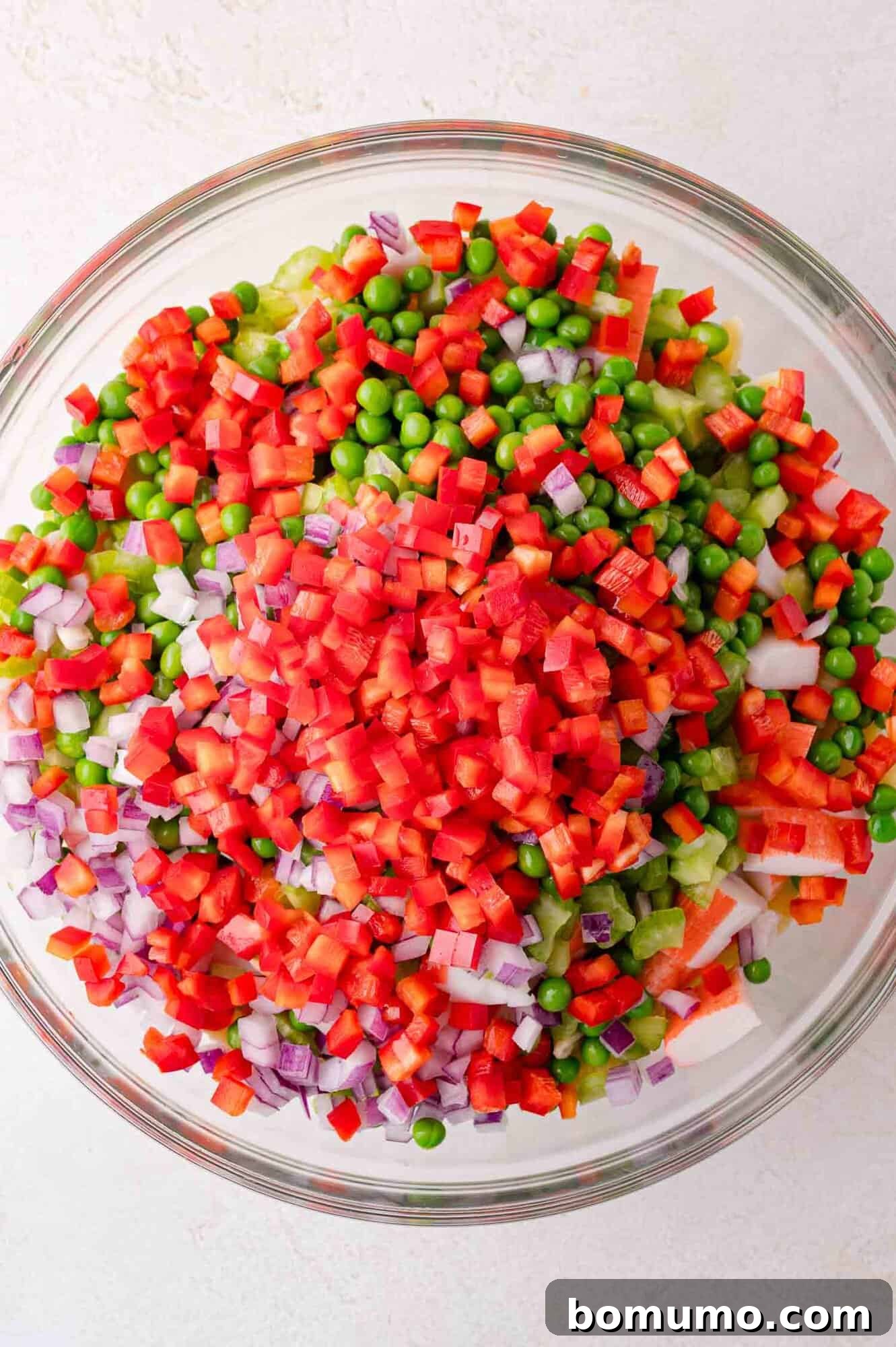 A vibrant glass bowl brimming with diced red bell peppers, bright green peas, chopped red onions, and a colorful mix of other fresh vegetables, ready for the pasta salad.