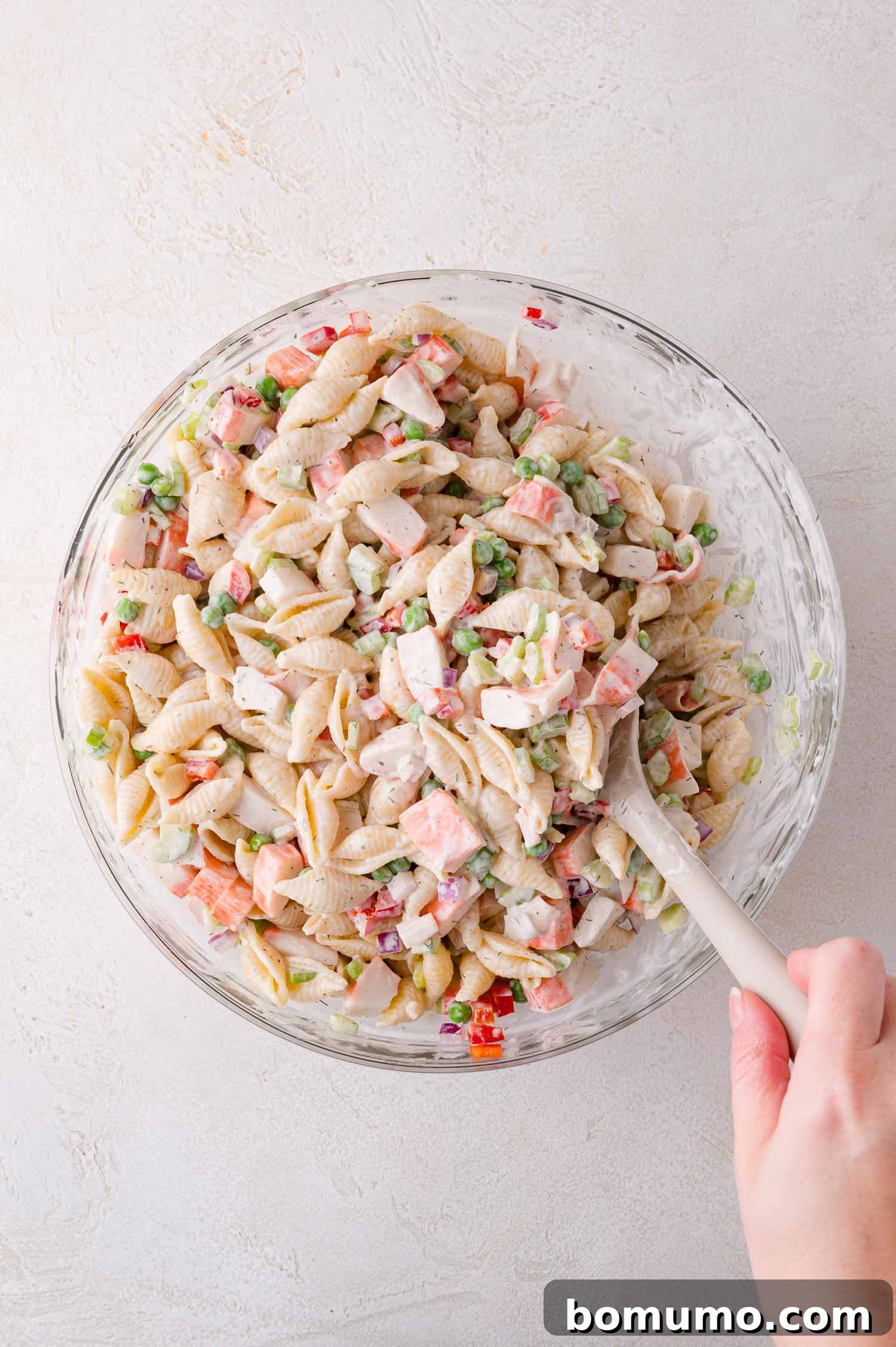 A hand gently stirs a glass bowl filled with a creamy pasta salad, featuring shell pasta, colorful chopped vegetables, and a rich dressing, demonstrating the mixing process.