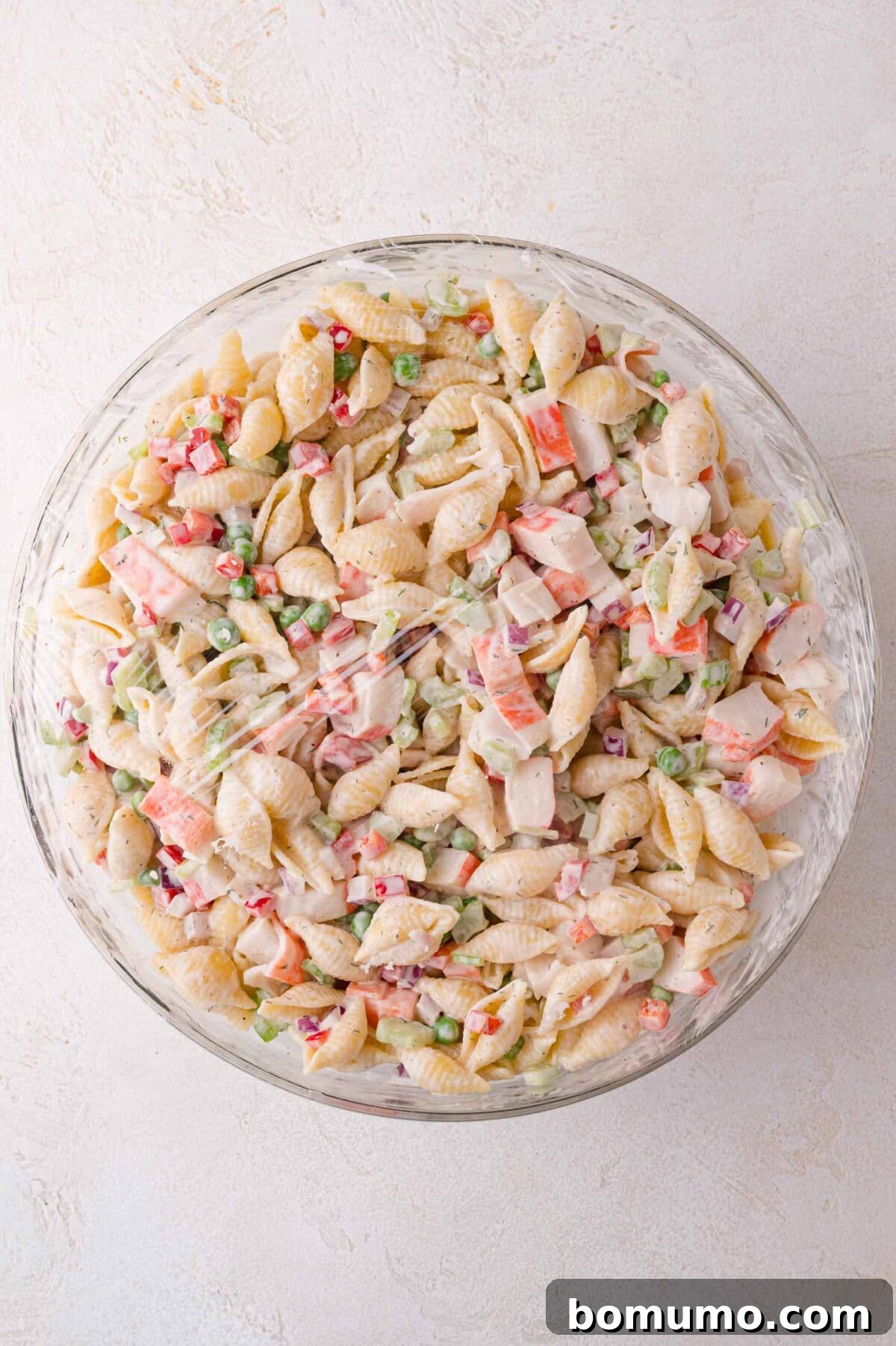 An inviting aerial view of a glass bowl filled with vibrant shell pasta salad, artfully mixed with imitation crab, crisp chopped vegetables, and a rich, creamy dressing, ready for serving.
