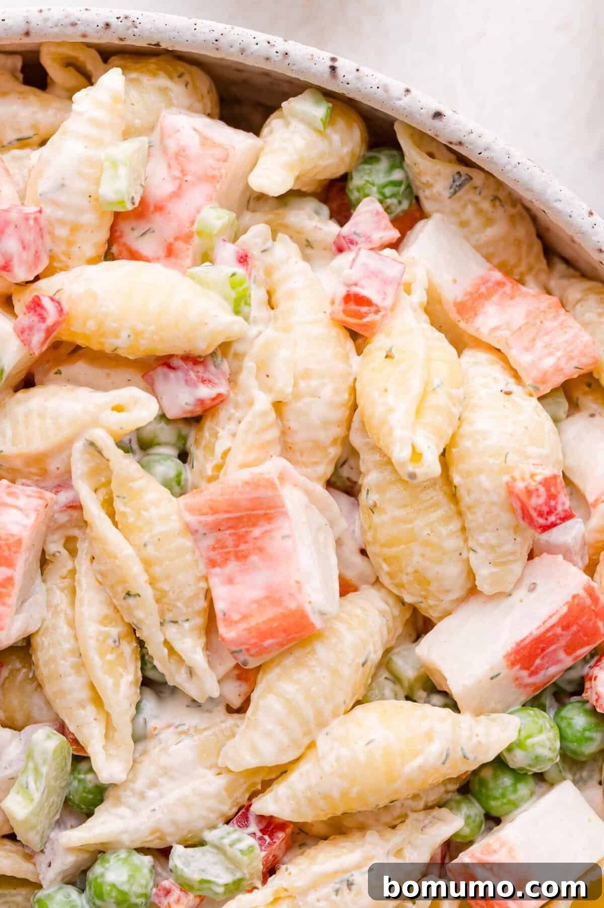 A close-up, appetizing view of perfectly cooked pasta shells intermingled with tender imitation crab meat, bright green peas, finely diced red bell pepper, and a generous coating of creamy dressing, highlighting the salad's delicious textures.