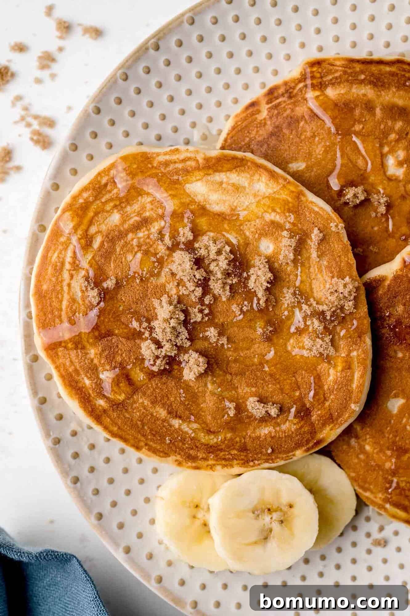 Molasses Sweetened Flapjacks 2 Two golden-brown pancakes richly topped with crumbled brown sugar and a generous drizzle of maple syrup, artfully arranged on a speckled white plate. Slices of fresh banana complement the dish, adding a touch of freshness.