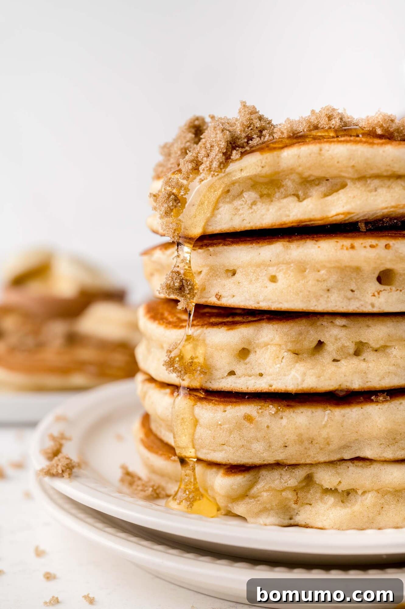 Molasses Sweetened Flapjacks 3 A beautifully stacked tower of five golden-brown pancakes, generously drizzled with golden syrup and garnished with a sprinkle of crumbled brown sugar, presented on an elegant white plate.