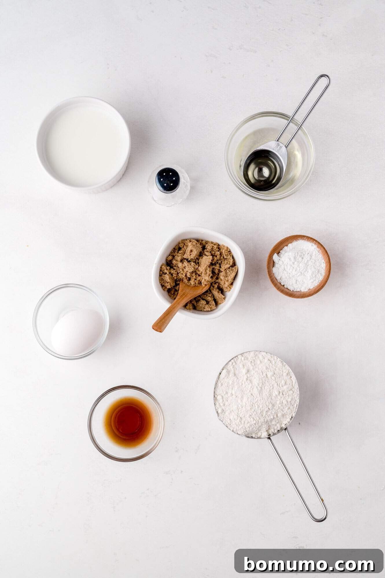 Molasses Sweetened Flapjacks 4 A collection of essential pancake ingredients neatly arranged on a pristine white surface. This includes a glass of milk, a single egg, a bowl of rich brown sugar, a bottle of pure vanilla extract, a mound of all-purpose flour, a small bowl of cooking oil, a pinch of salt, and a measuring spoon of baking soda, all prepared for mixing.