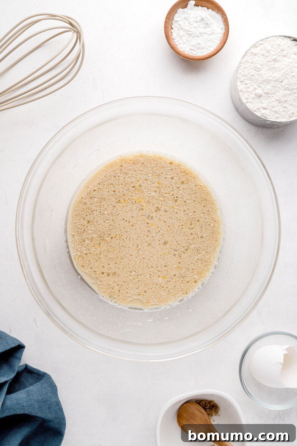 Molasses Sweetened Flapjacks 6 A mixing bowl filled with smooth, golden liquid batter takes center stage on a white surface. It is surrounded by essential baking tools: a whisk, a pile of flour, a small container of baking powder, and a bowl holding a spoon. A soft napkin lies nearby, ready for use.