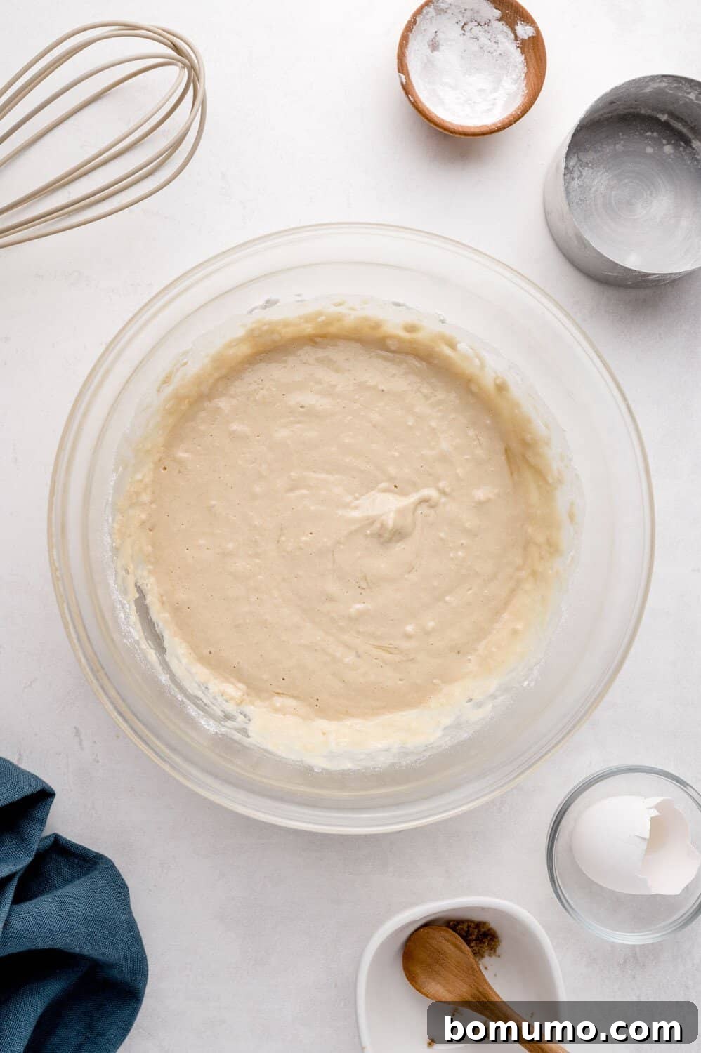 Molasses Sweetened Flapjacks 7 A clear glass bowl containing a rich, mixed cake batter rests on a clean white surface. Surrounding the bowl are an array of baking essentials: a whisk, fresh eggs, small ingredient bowls, and a soft blue cloth, all suggesting an active cooking session.