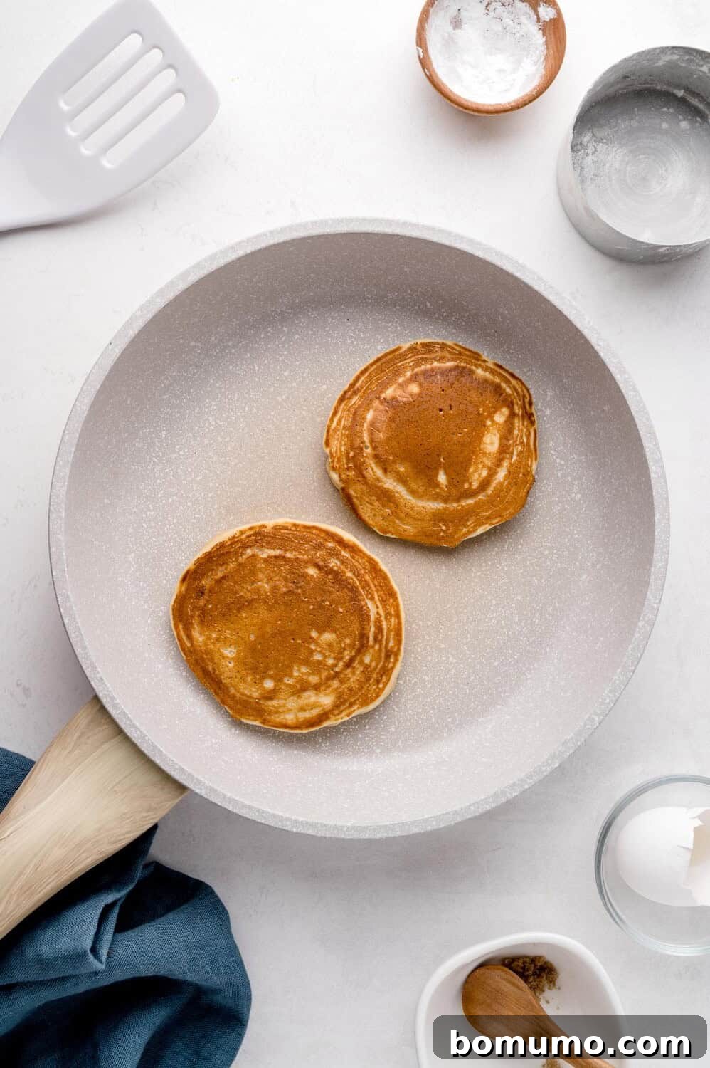 Molasses Sweetened Flapjacks 8 Two perfectly round pancakes sizzle gently in a sleek gray frying pan, radiating warmth. The pan is surrounded by an assortment of baking ingredients and various utensils, all neatly arranged on a clean white surface, suggesting a delightful breakfast preparation.