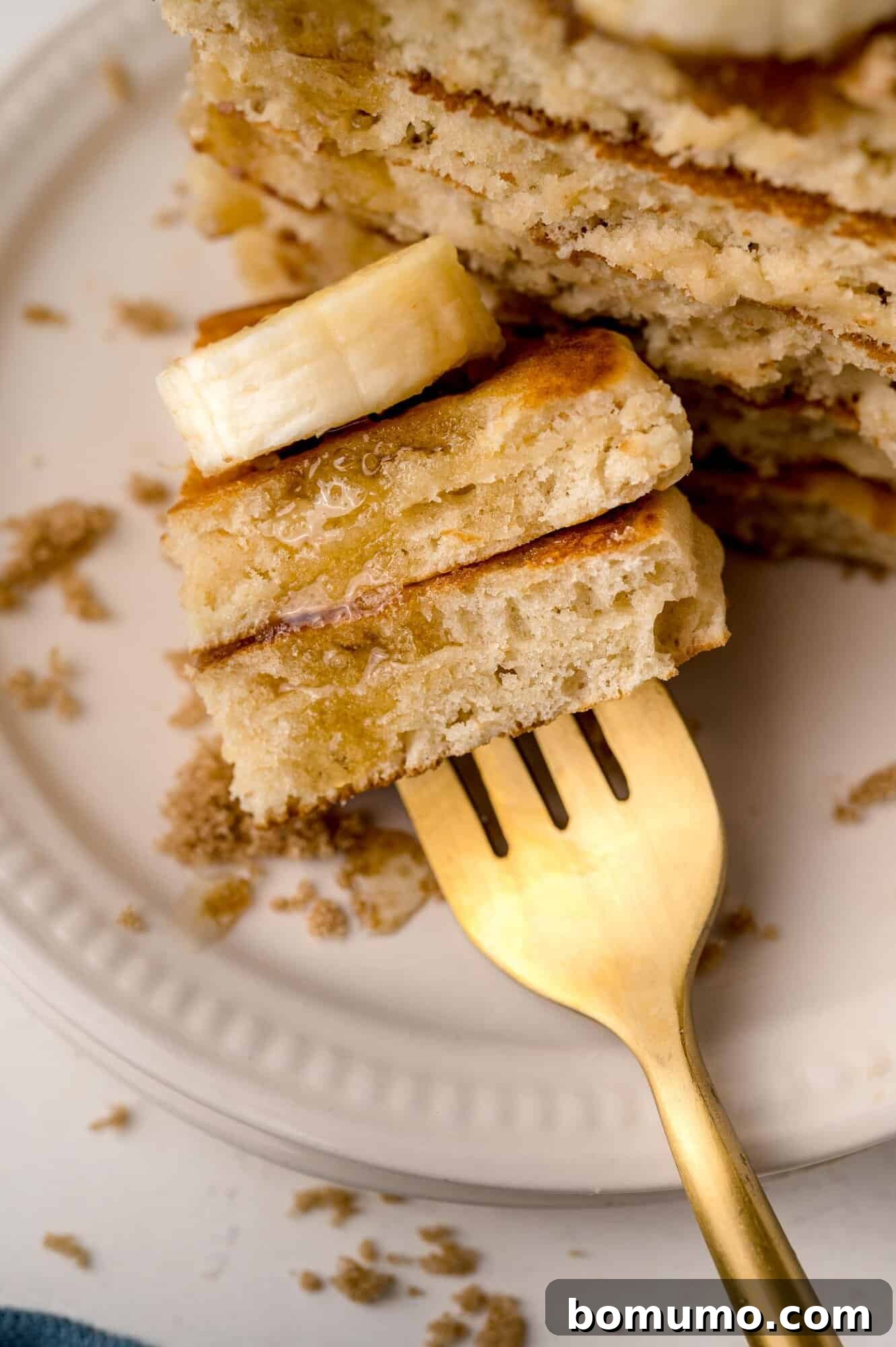 Molasses Sweetened Flapjacks 9 A fork lifts a delectable bite of pancake, garnished with banana slices and a generous drizzle of syrup, from a white plate. The plate itself is artfully sprinkled with brown sugar, enhancing the visual appeal.
