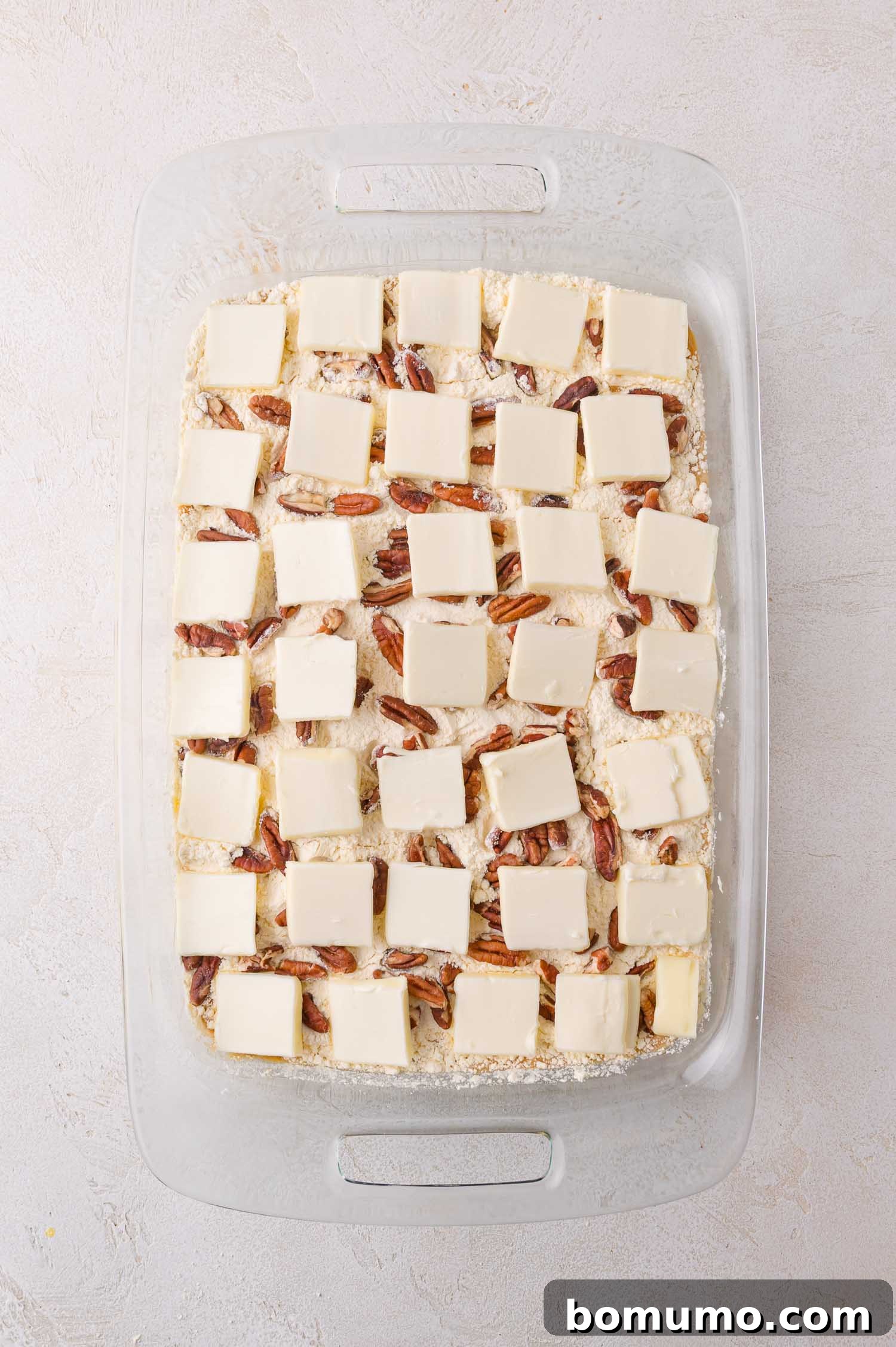 A glass baking dish containing the unbaked dump cake now features neatly arranged, evenly spaced slices of butter over the pecans and crumbly cake mixture, poised for baking and promising a golden, crispy topping.