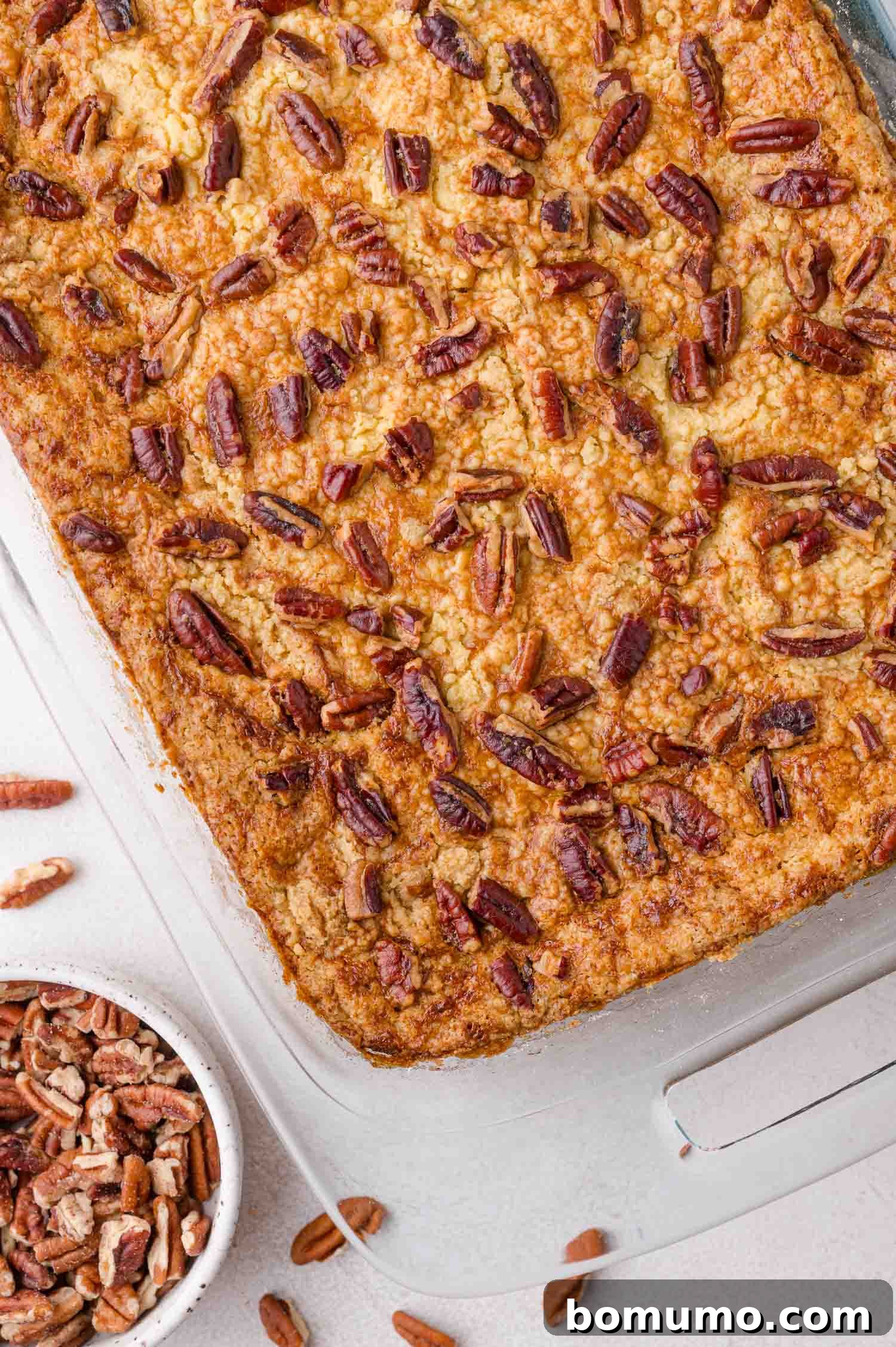 A glass baking dish showcases a freshly baked pumpkin dump cake, its top adorned with golden-brown chopped pecans. A small bowl of additional pecans sits nearby on a light surface, suggesting a finishing touch or extra crunch.