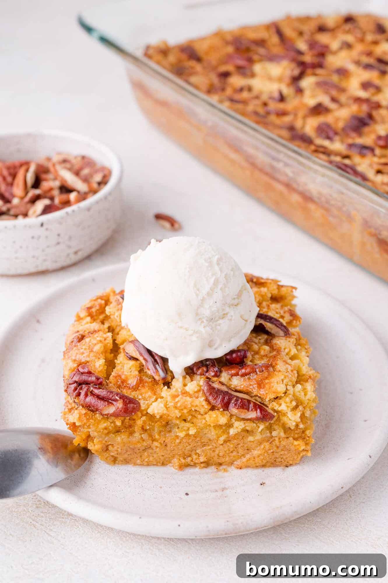 A generous slice of pumpkin dessert, elegantly topped with crunchy pecans and a creamy scoop of vanilla ice cream, rests on a plate with a spoon at its side. In the soft background, the full baking dish of the delicious dessert awaits, ready for more servings.