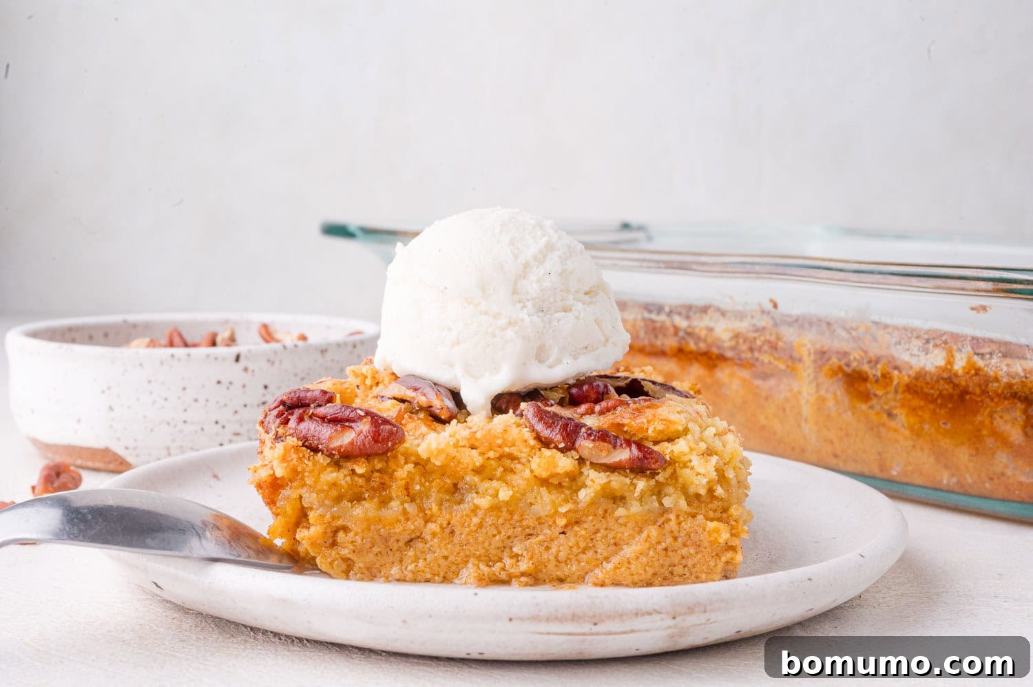 A perfectly sliced serving of pumpkin dessert, adorned with crunchy pecans and a dollop of melting vanilla ice cream, presented on a pristine white plate. A fork rests beside it, with a full baking dish visible in the soft background, ready for more servings.