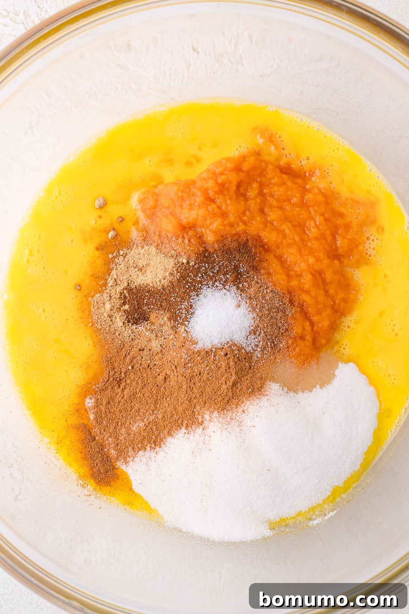A clear glass mixing bowl holds the initial ingredients for the pumpkin custard: beaten eggs, creamy pumpkin puree, sparkling granulated sugar, a pinch of salt, and a medley of warm spices, all awaiting a final whisk to blend their flavors.