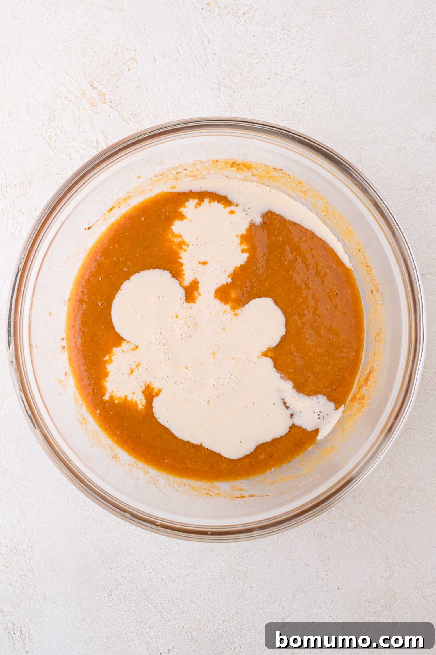 A light-colored surface hosts a clear glass bowl, showcasing a rich orange pumpkin puree with evaporated milk being partially stirred in, creating a marbled effect as the two liquids begin to combine for the custard base.