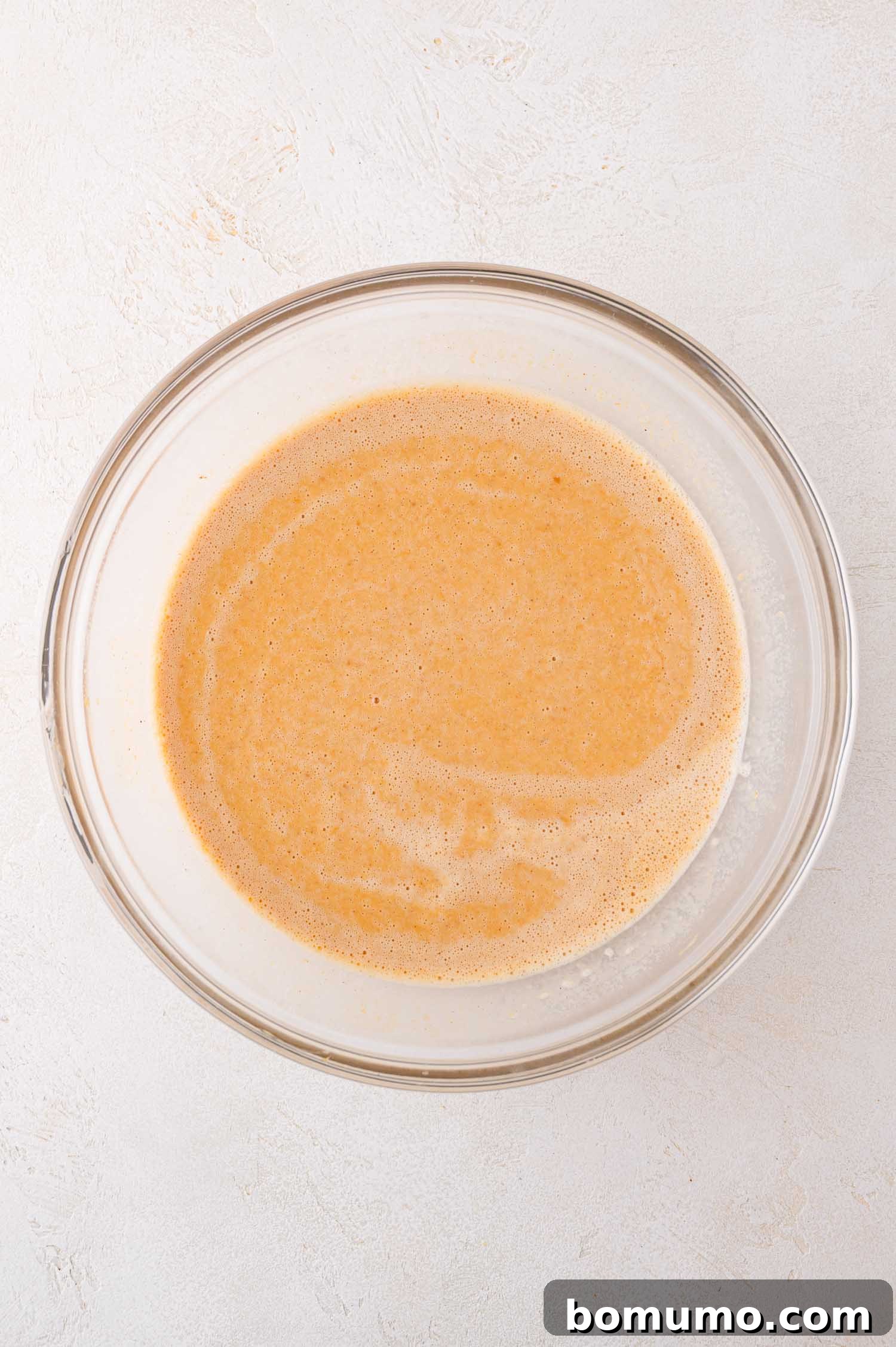 A clear glass mixing bowl, filled with a smooth, light brown pumpkin mixture, rests on a subtly textured white surface, indicating the custard base is perfectly prepared and ready for the next step in the dump cake assembly.