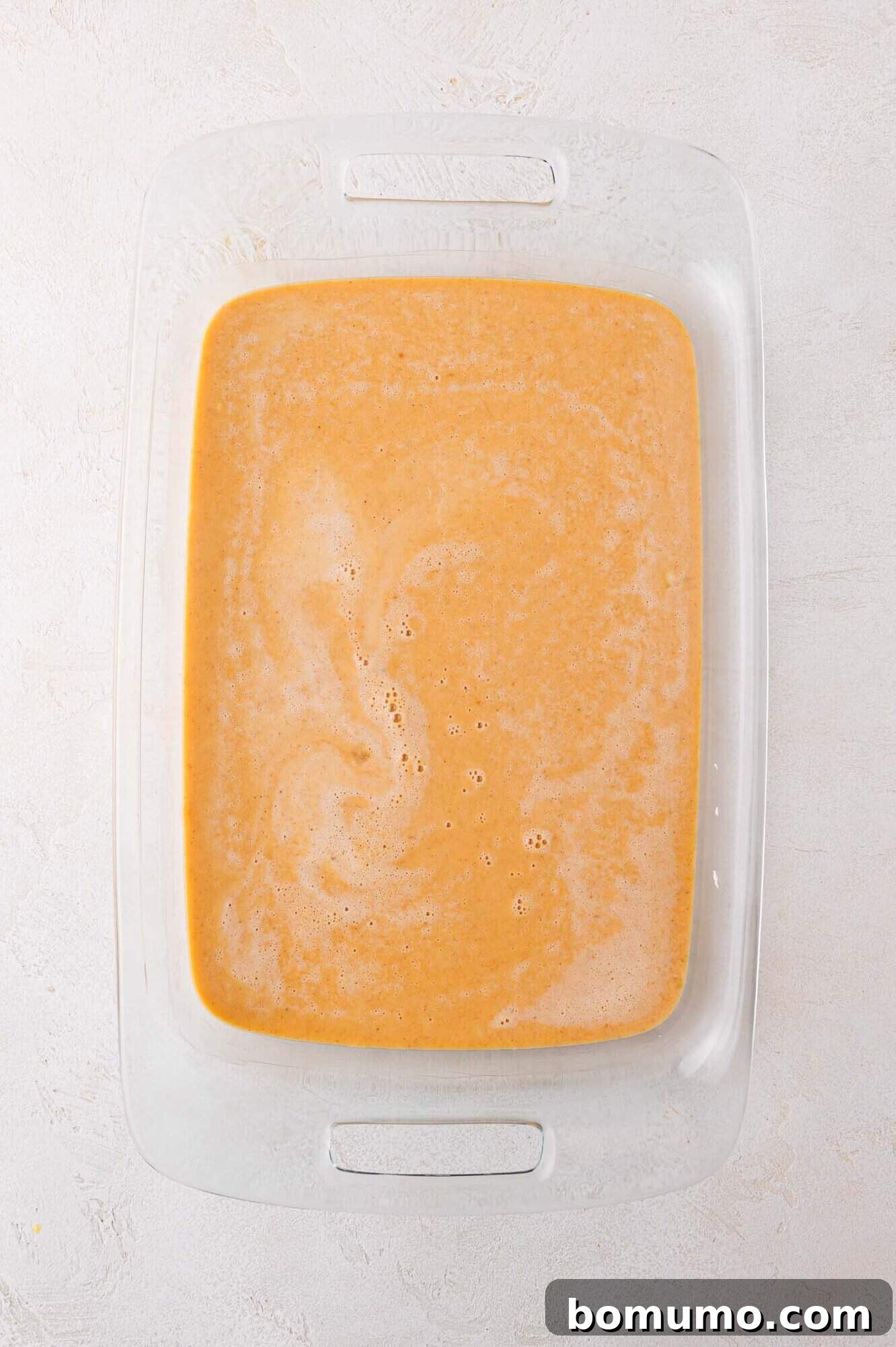 A clear glass baking dish now holds a smooth, glistening layer of orange-brown liquid, the meticulously prepared pumpkin filling, resting on a softly textured light surface, awaiting the next layers of the dump cake.