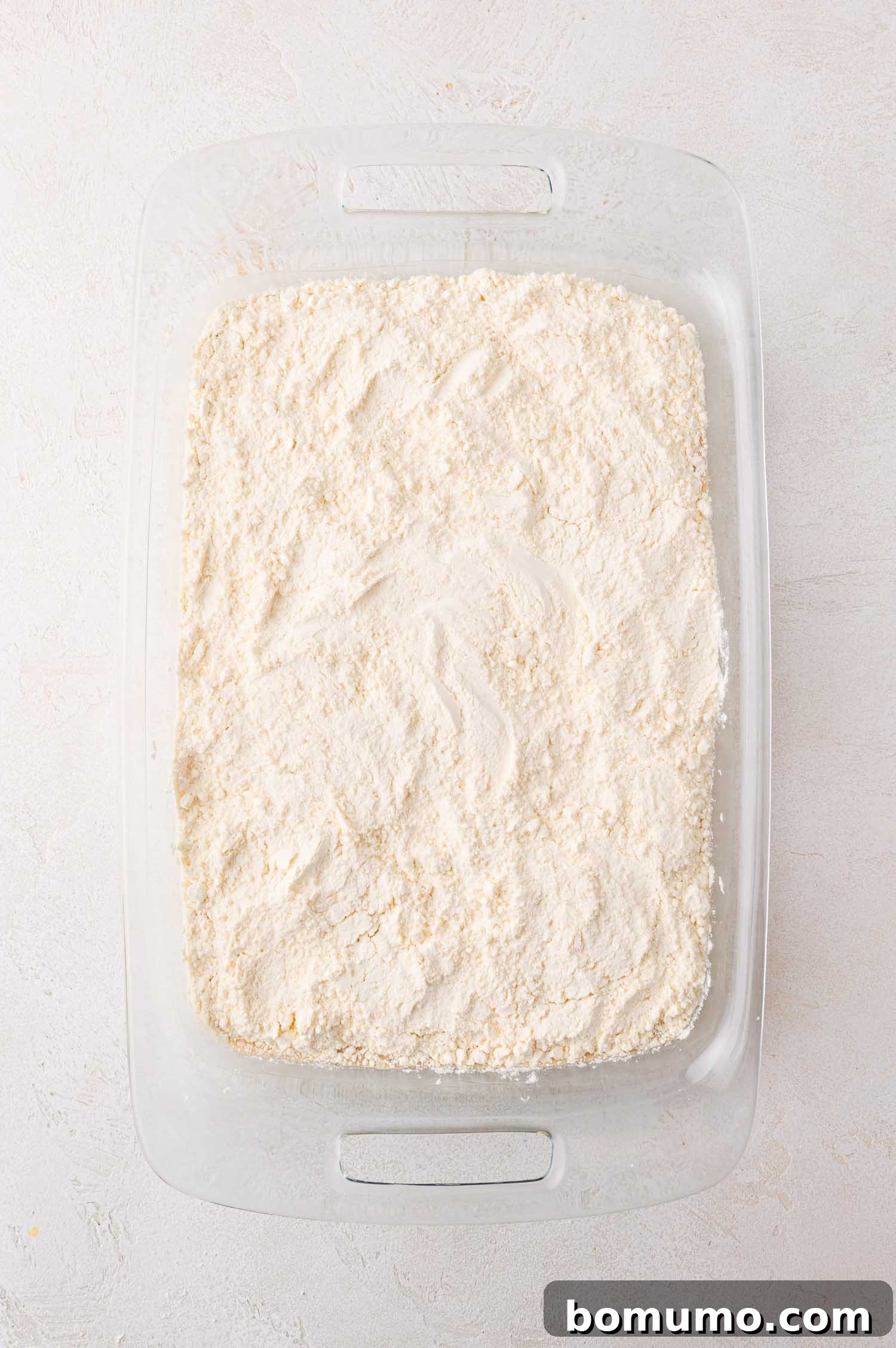 A rectangular glass baking dish is now evenly covered with a pristine white layer of dry, crumbly cake mix, meticulously spread over the pumpkin filling, ready for the next additions on a light surface.