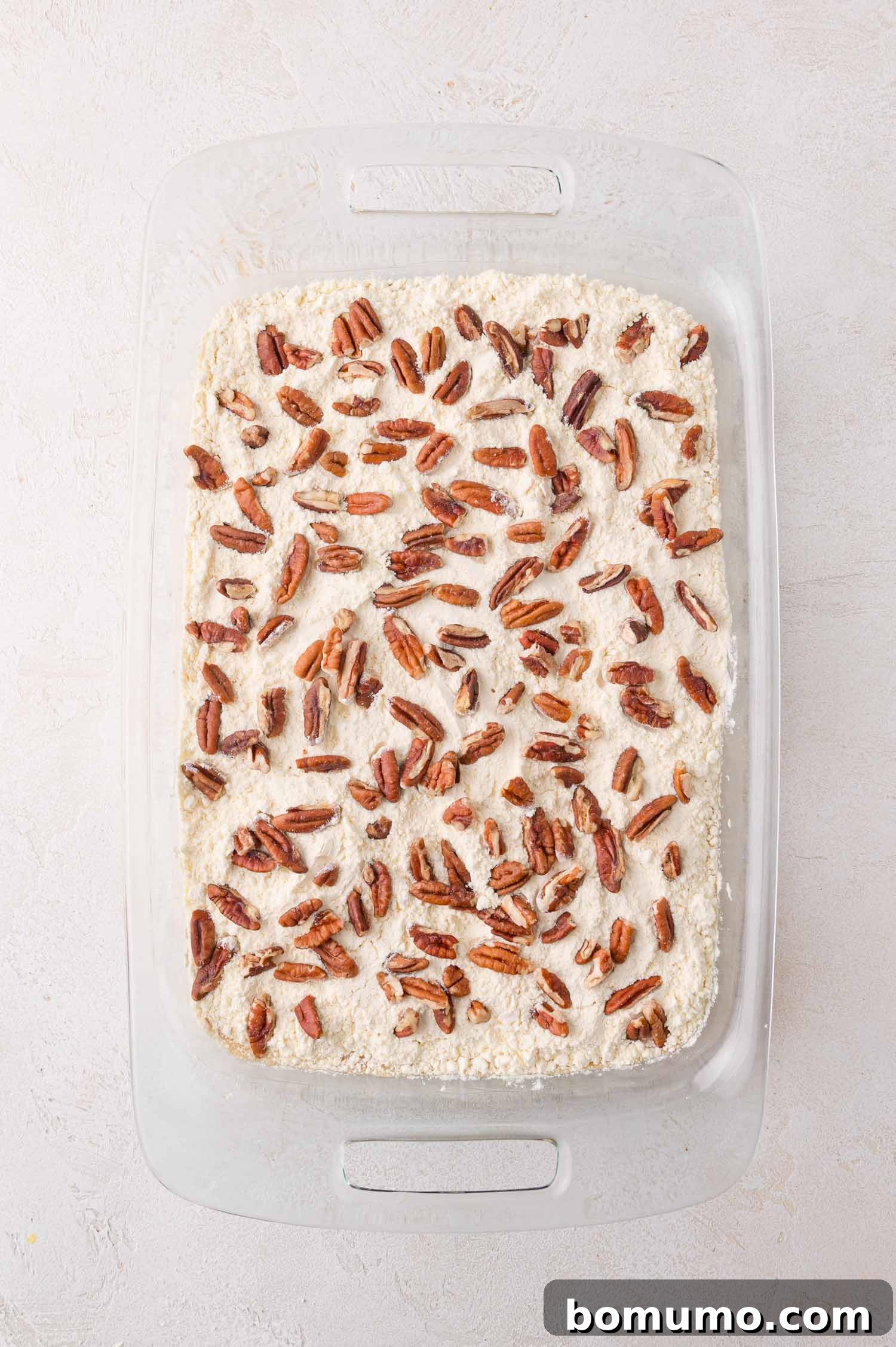 An unbaked pumpkin dump cake dessert fills a glass baking dish, now beautifully topped with a generous scattering of chopped pecans over the dry cake mix, creating an inviting texture contrast on a light-colored surface.
