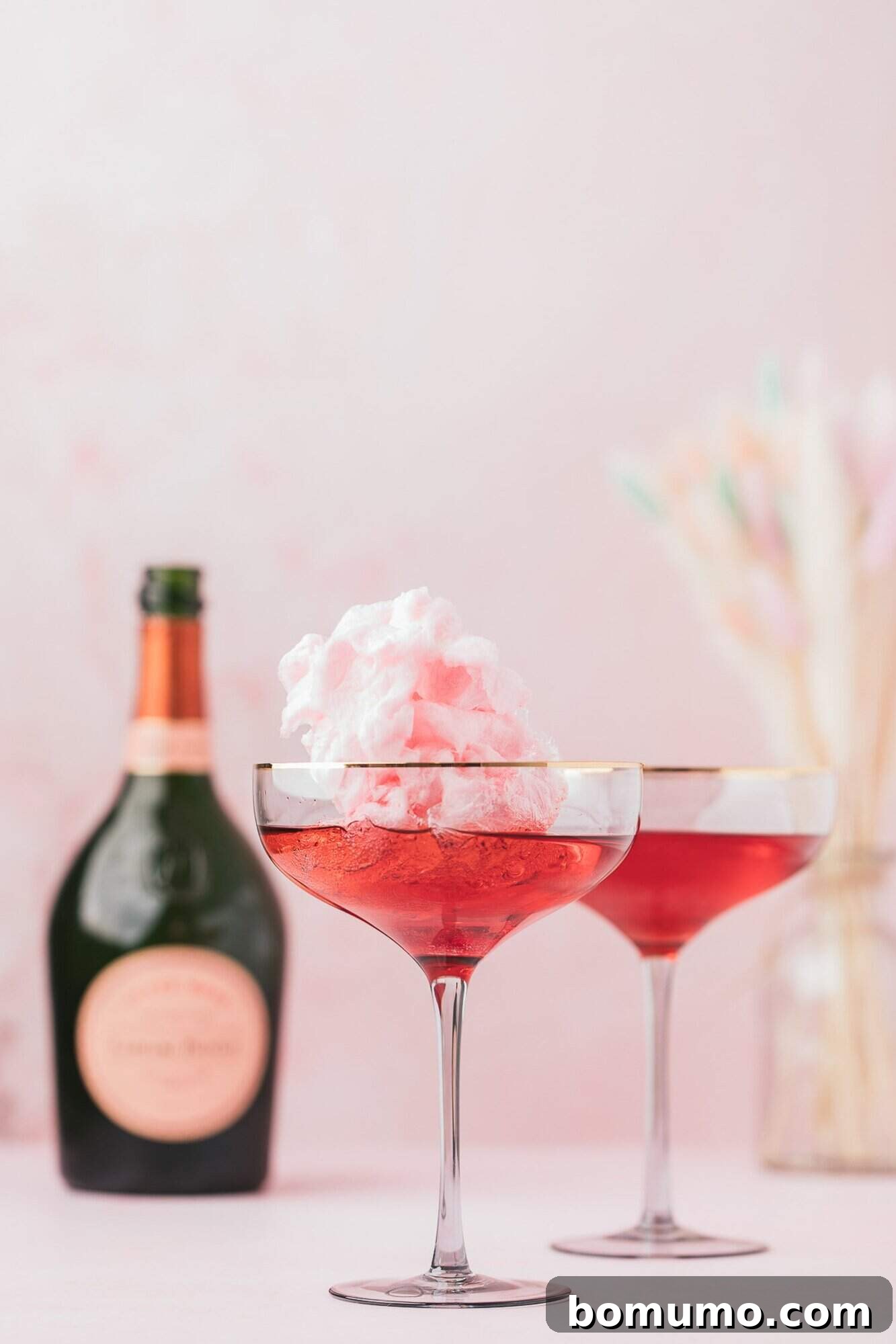 Two glasses of sparkling pink cotton candy cocktails garnished with fluffy pink cotton candy, set against a soft pink background with a bottle of sparkling wine.