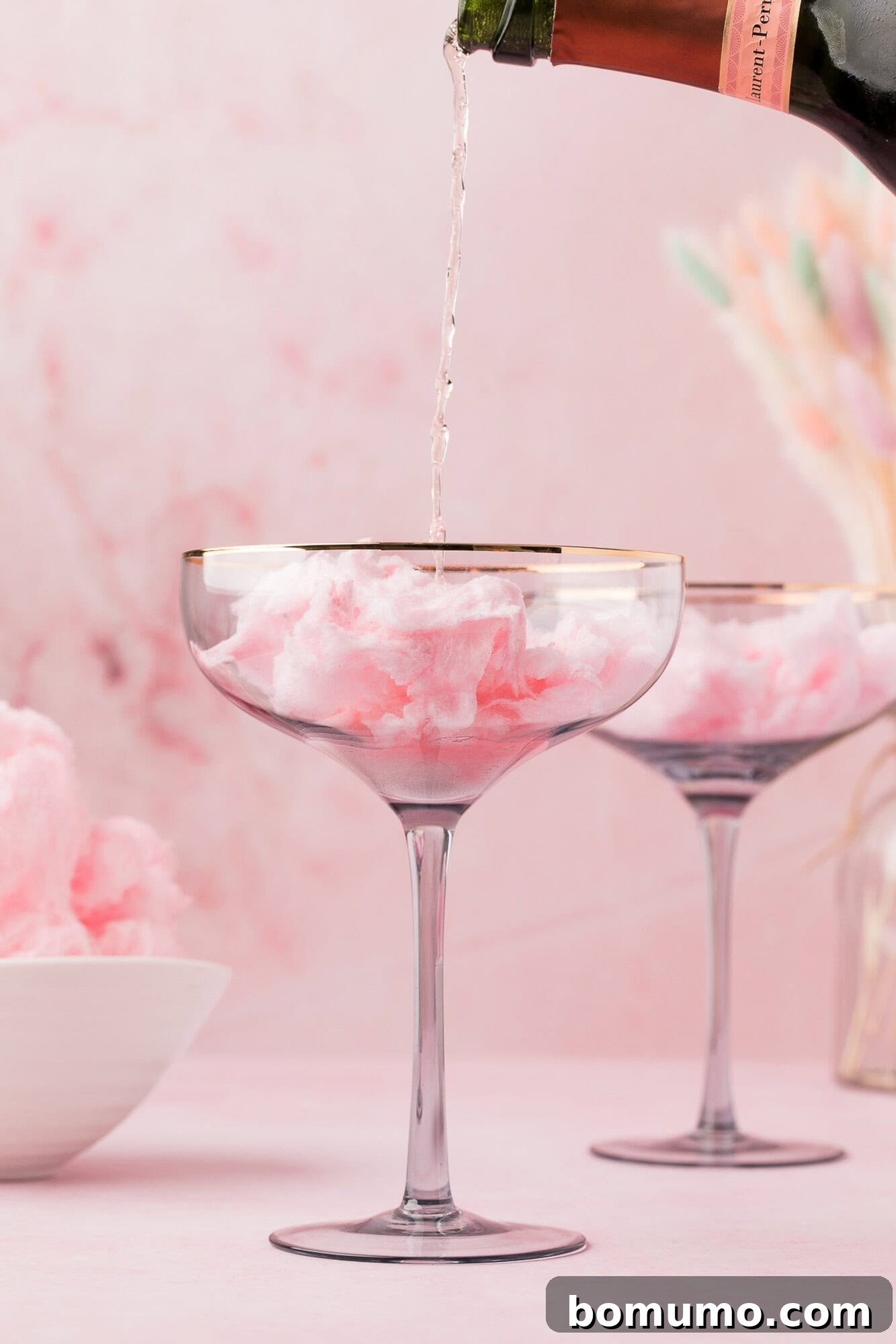 Champagne being poured into a glass filled with pink cotton candy, with another similar glass and a bowl of cotton candy in the background, set against a pink backdrop.