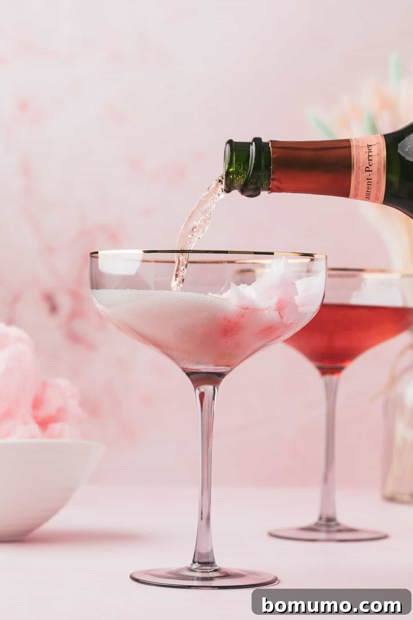 Sparkling wine being poured into a glass with cotton candy inside, with another similar drink and a bowl of cotton candy in the background.