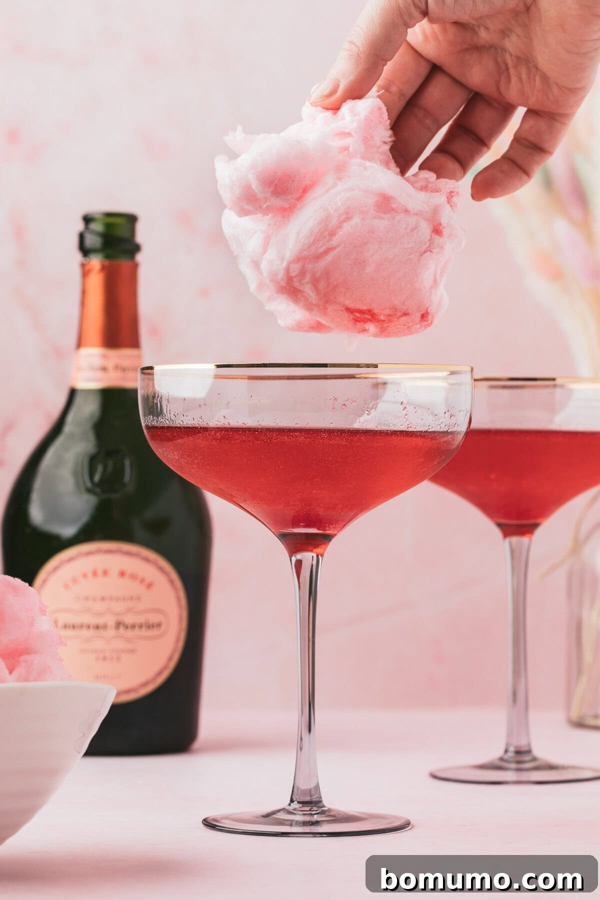 A hand places a fluffy cloud of pink cotton candy atop a glass of pink sparkling cocktail. A bottle of sparkling rosé and another garnished cocktail are visible in the soft pink background.