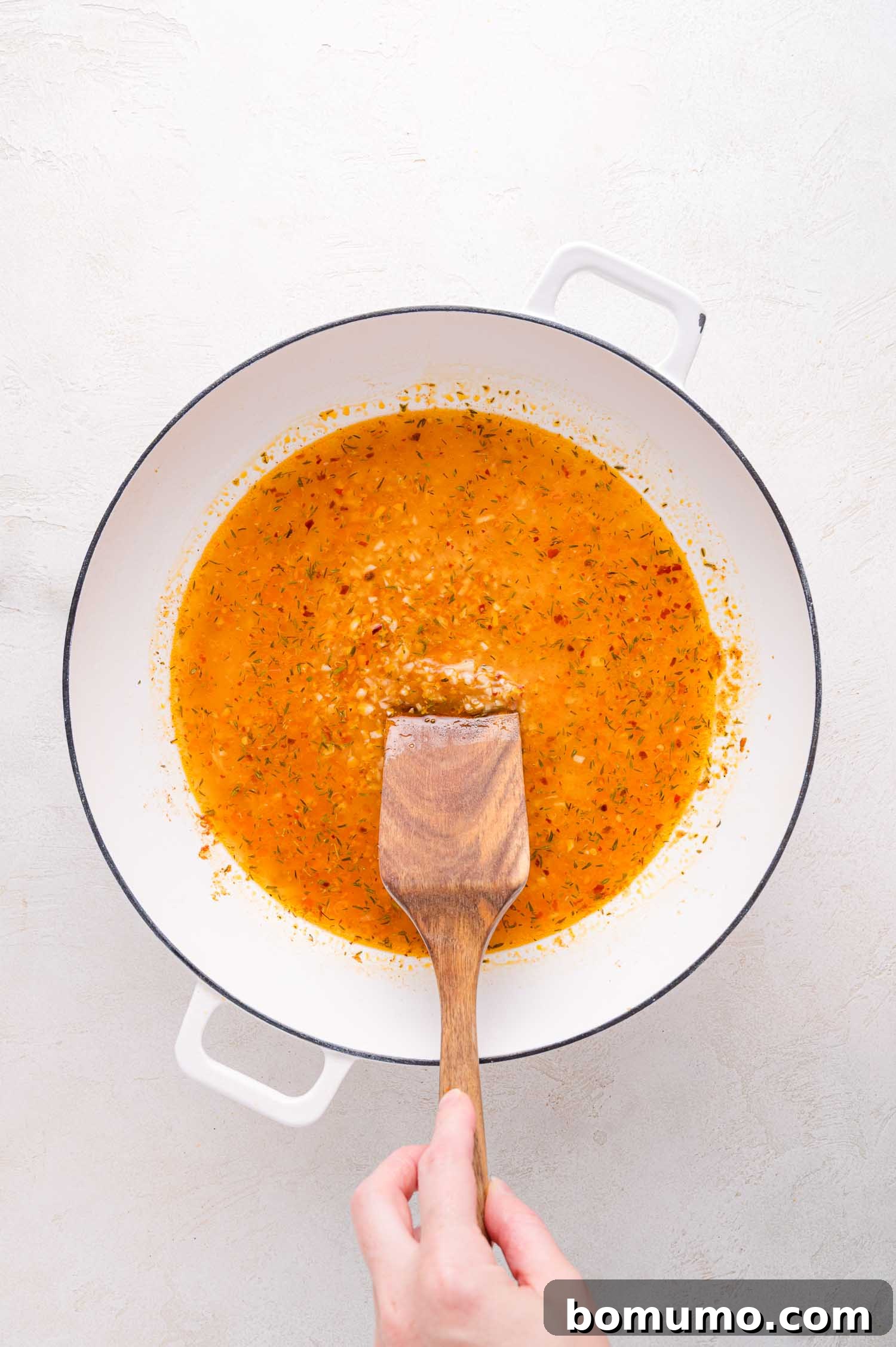 A hand expertly stirs a rich, orange-colored sauce infused with fragrant herbs within a pristine white pot, utilizing a rustic wooden spatula, demonstrating the careful blending of flavors for the Lemon Butter Chicken.