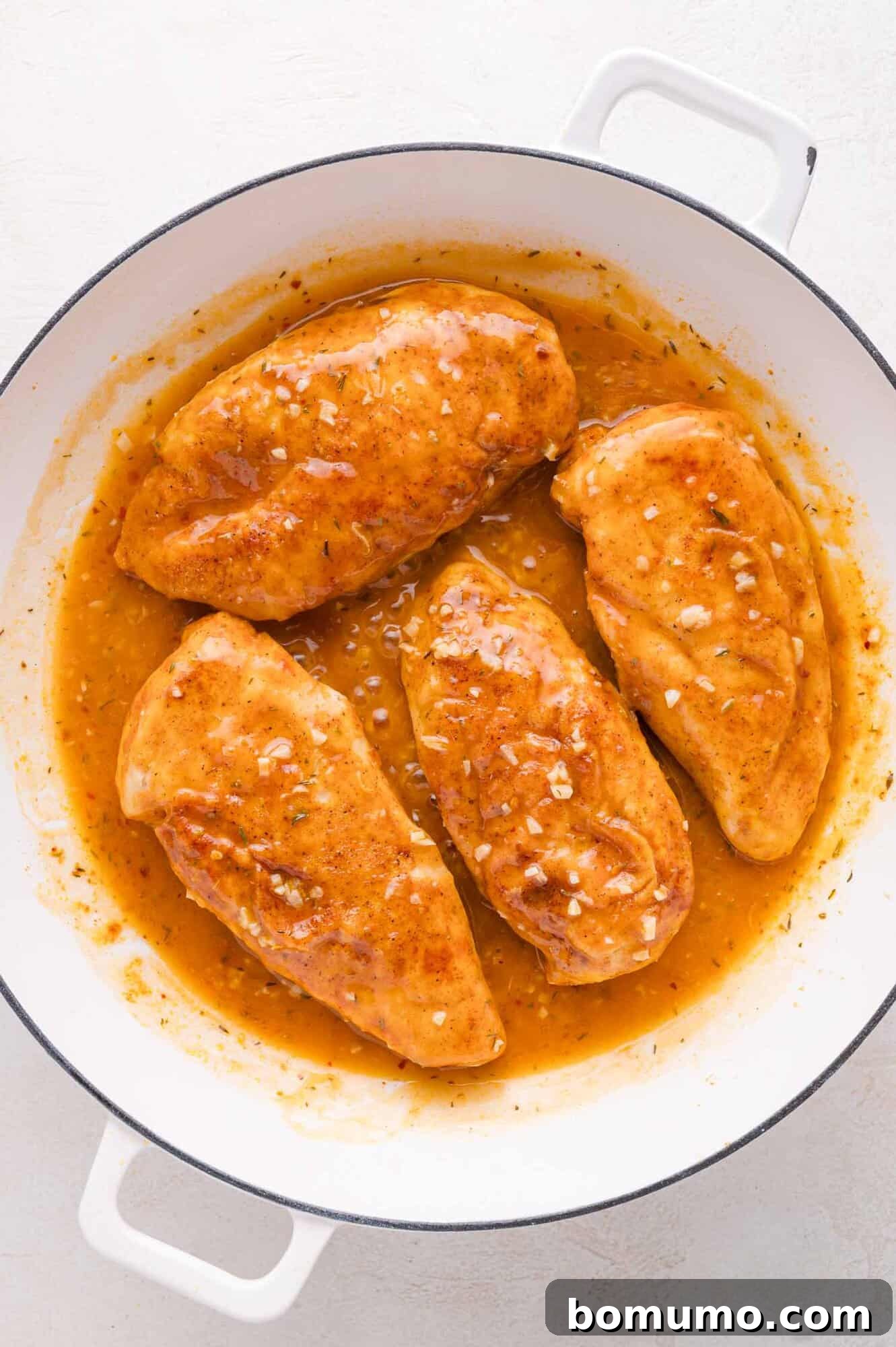 Four perfectly seasoned chicken breasts are delicately simmering in a luscious, light brown sauce within a crisp white skillet, with visible specks of finely diced garlic distributed throughout the rich sauce, infusing every bite with exquisite flavor.