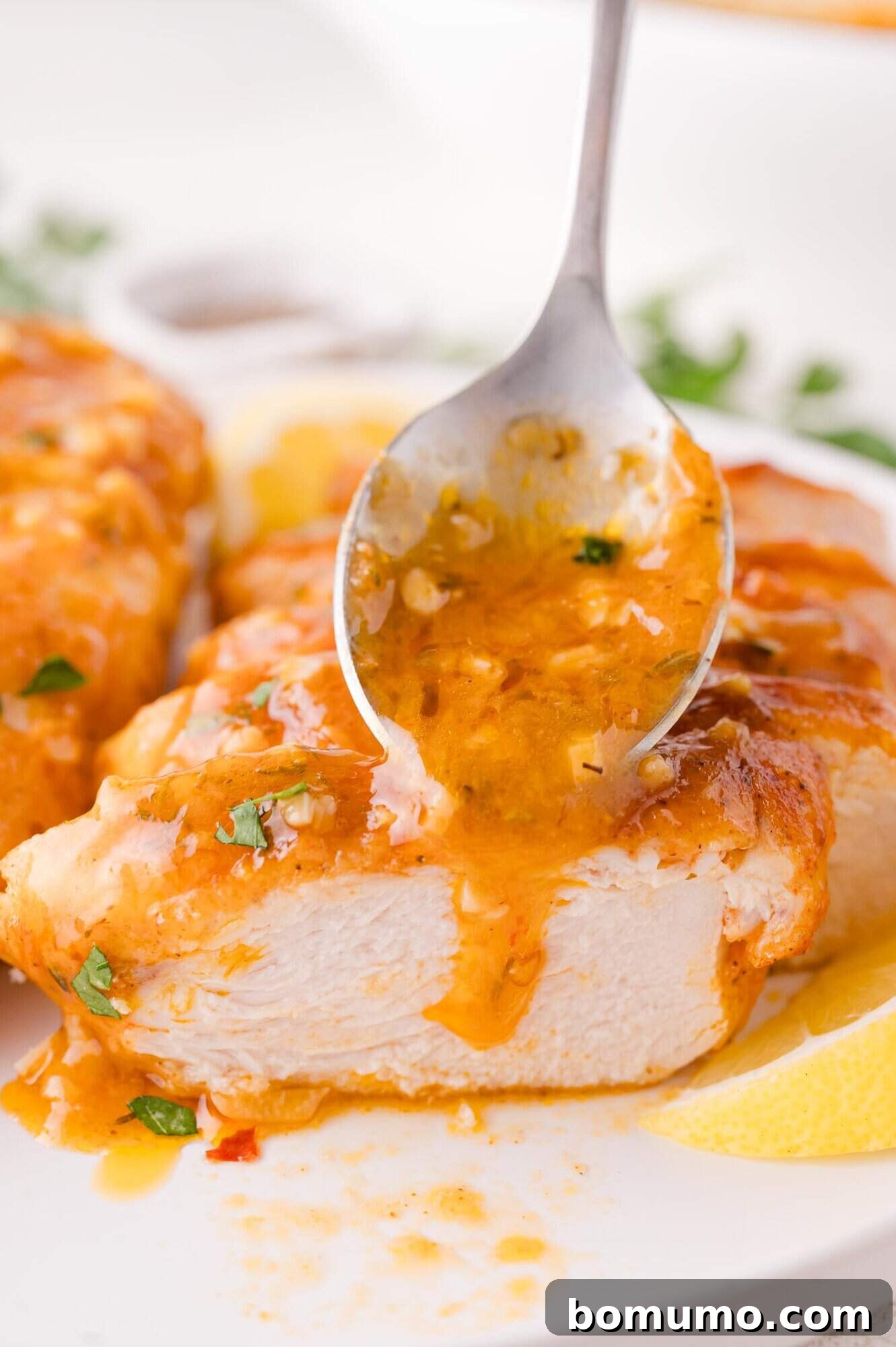 A elegant spoon gracefully pours a vibrant orange sauce over a perfectly sliced piece of cooked chicken breast, which is artfully garnished with fresh herbs, resting on a pristine white plate alongside a bright lemon wedge, creating a visually stunning and delicious presentation.