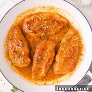 Four perfectly cooked chicken breasts nestled in a gleaming skillet, generously coated in a thick, vibrant orange-brown garlic butter sauce and beautifully garnished with fresh chopped herbs.