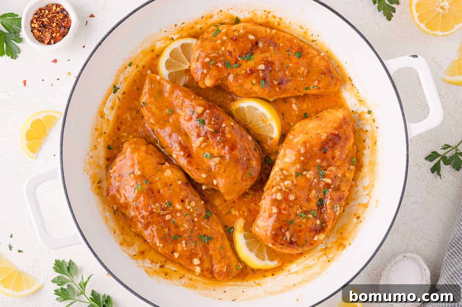 Four perfectly cooked chicken breasts nestled in a gleaming white skillet, bathed in a rich, golden lemon-garlic sauce. The dish is elegantly garnished with vibrant lemon slices and a sprinkle of fresh, chopped herbs, enhancing both its visual appeal and aromatic charm. This pan-cooked masterpiece promises a succulent and intensely flavorful experience, reminiscent of a gourmet restaurant entrée, all while being wonderfully easy to prepare at home.
