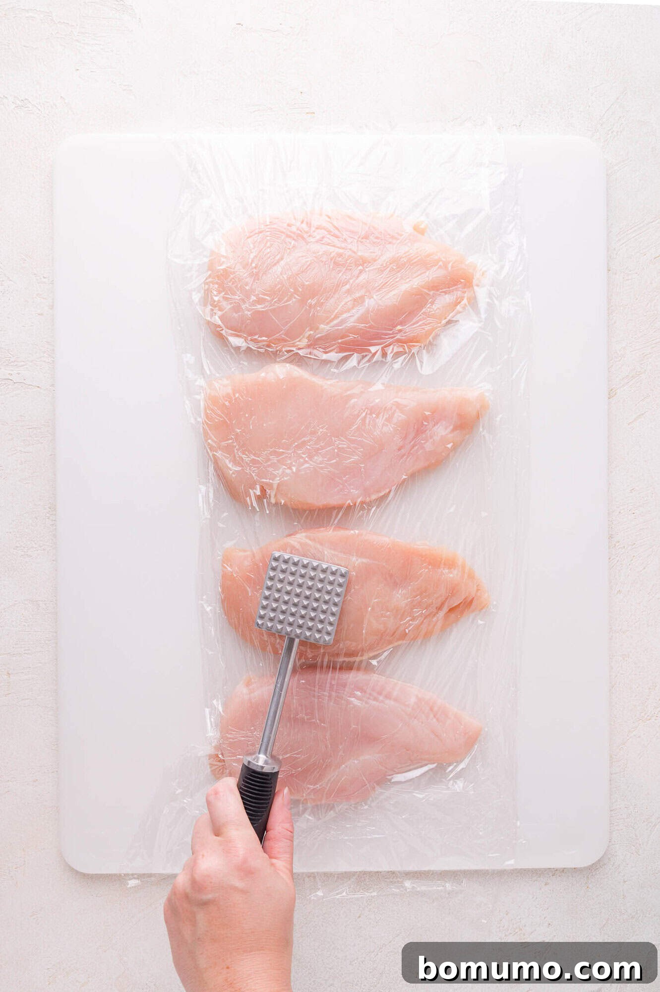 A close-up view of three raw chicken breasts neatly arranged on a cutting board, covered with a layer of clear plastic wrap. A hand firmly holds a meat mallet poised directly above the chicken, ready to tenderize and flatten it to an even thickness, ensuring perfect cooking for the Lemon Butter Chicken.