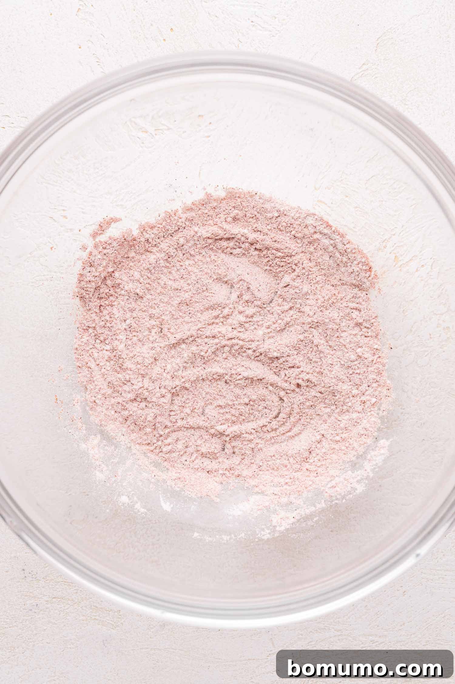 A transparent glass bowl neatly filled with a finely ground, light pinkish-brown spice mixture, composed of flour, salt, paprika, and pepper, resting on a clean white textured surface, showcasing the perfectly blended dredging ingredients for the chicken.