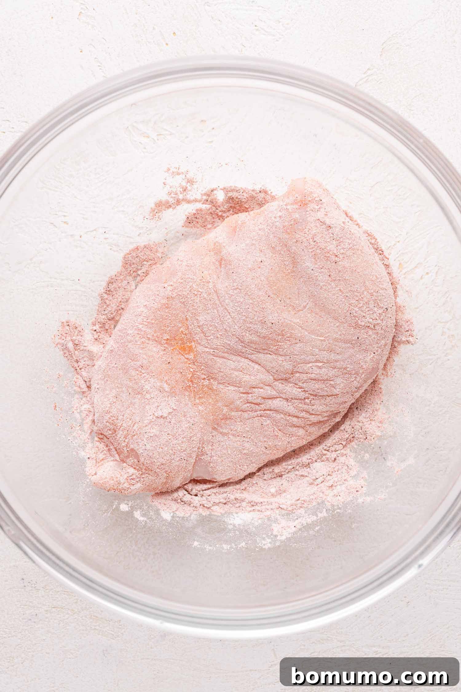 A single raw chicken breast, thoroughly coated with a delicate, pink-hued seasoning blend, rests within a clear glass bowl, positioned on a smooth, light-colored surface, illustrating the successful dredging process of the chicken for the recipe.