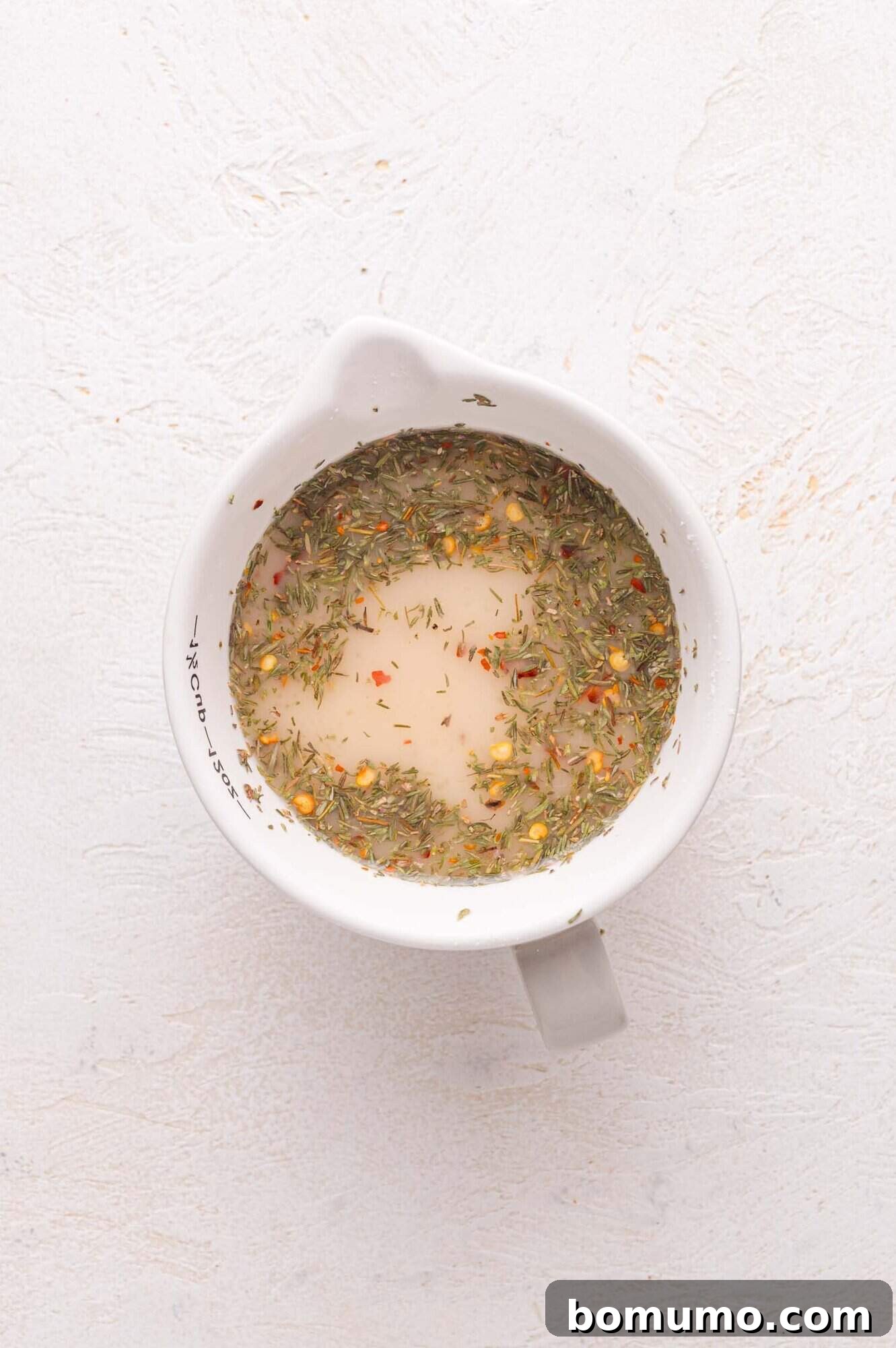 A pristine white measuring cup, viewed from above, holds a vibrant, herb-flecked sauce mixture, comprising chicken broth, lemon juice, cornstarch, lemon zest, thyme, and red pepper flakes, artfully arranged on a light textured surface, awaiting its turn in the skillet.
