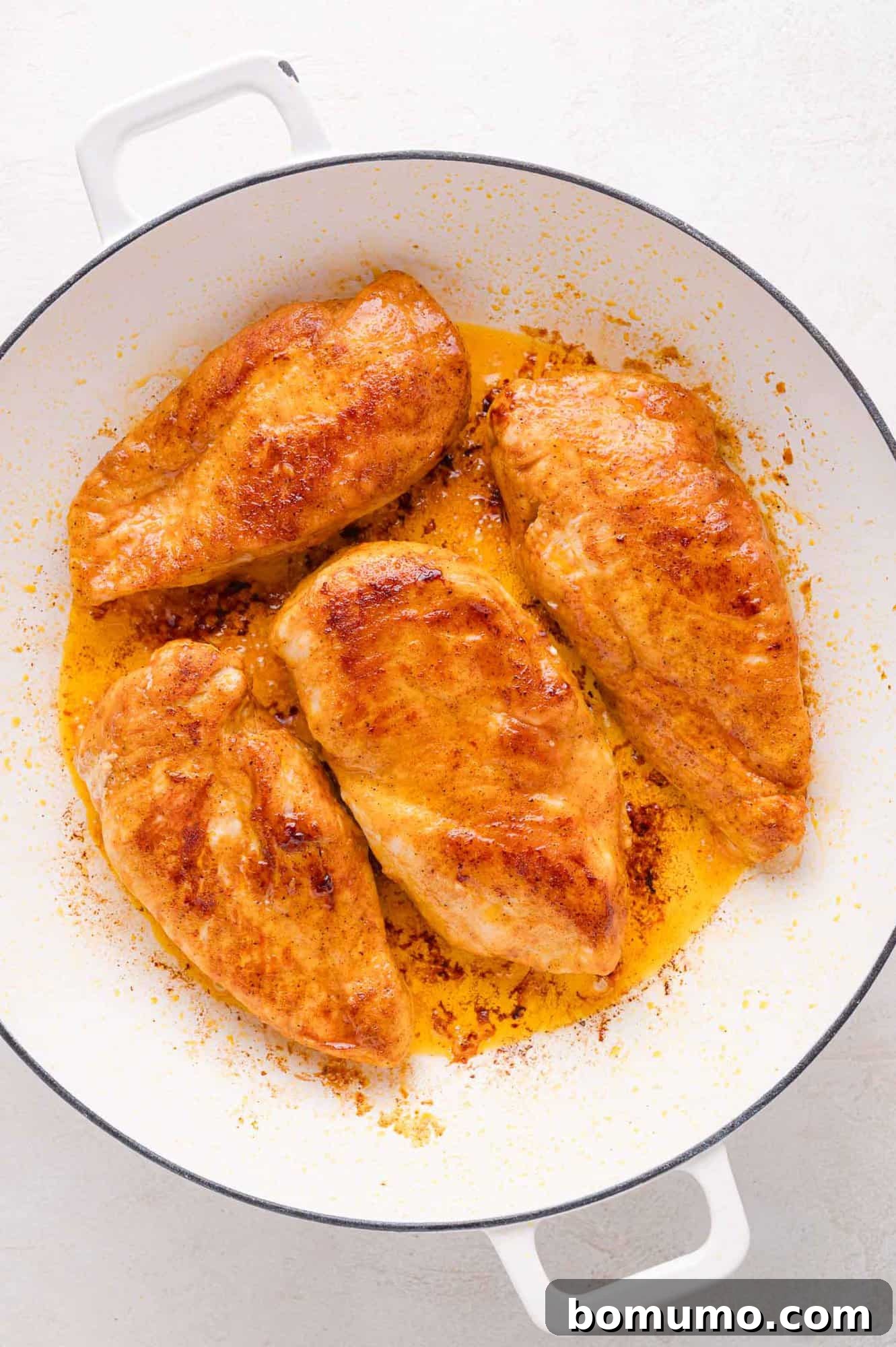 Four perfectly cooked, seasoned chicken breasts are meticulously arranged in a clean white skillet, showcasing their appealing browning and visible, savory juices, indicating they are ready for the next step in the Lemon Butter Chicken recipe.