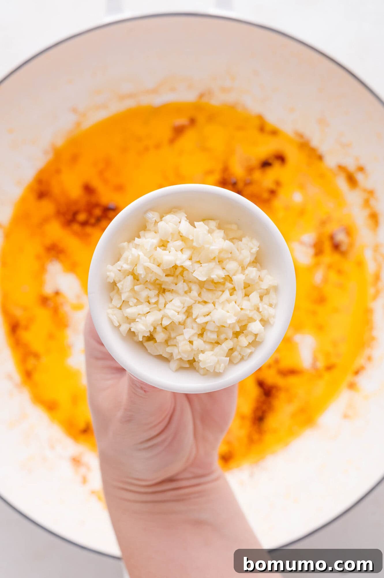 A skilled hand gently sprinkles a bowl of finely minced garlic directly above a warm pan, where olive oil and a delicate blend of spices are already simmering, signaling the commencement of the flavorful sauce preparation.