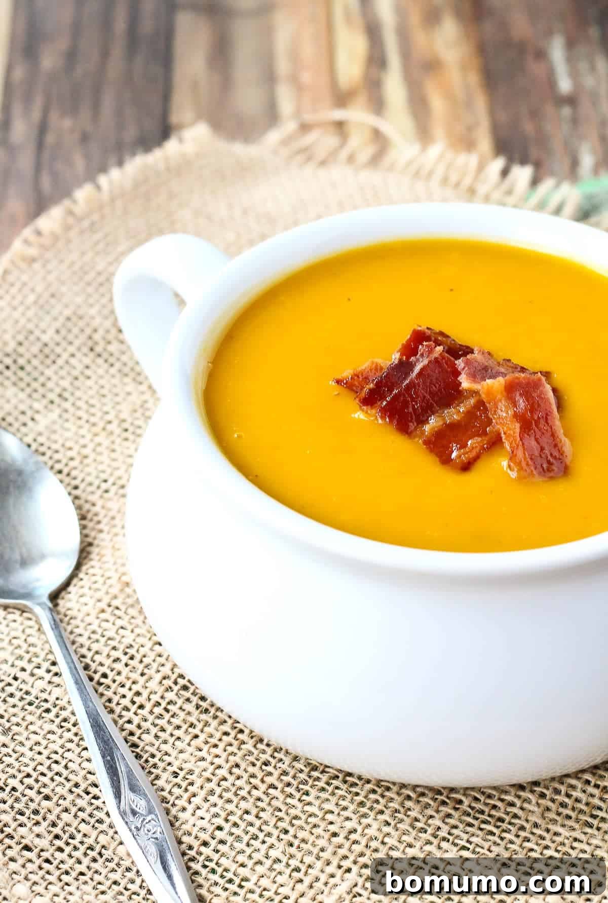 A vibrant orange butternut squash soup in a pristine white bowl, garnished generously with golden, crispy bacon bits. The backdrop suggests a cozy, inviting kitchen setting.