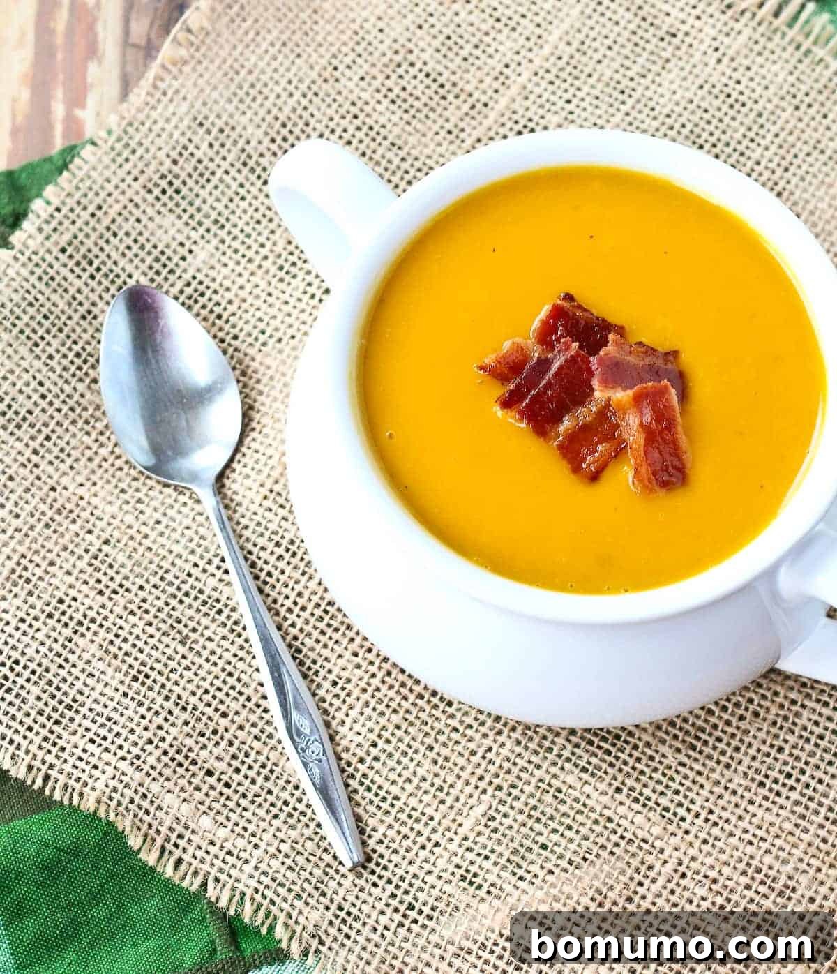 An inviting overhead shot of a white bowl filled with creamy butternut squash soup, garnished with vibrant green herbs and crispy, crumbled bacon pieces. The texture of the soup is smooth and rich.