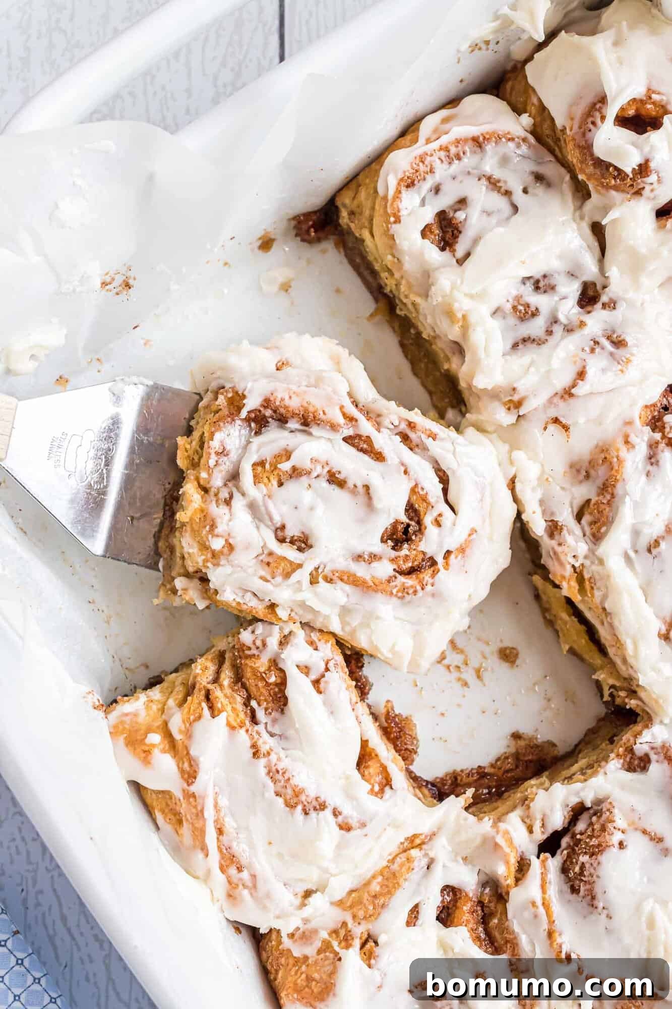 Quick Cinnamon Crescent Swirls 2 A spatula lifts a frosted cinnamon roll from a baking dish containing several iced cinnamon rolls, ready to be enjoyed.